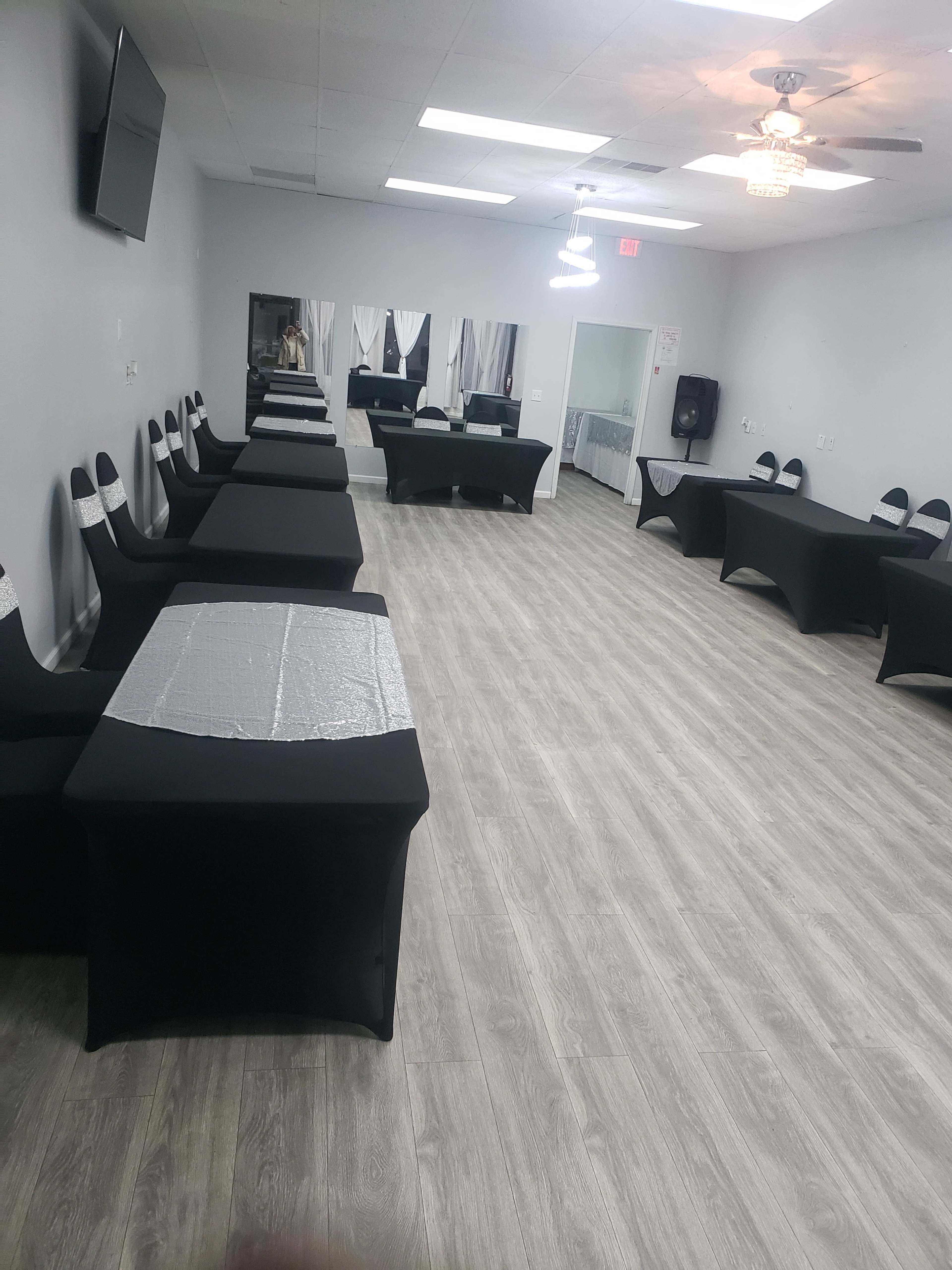 A spacious event room with black tables and chairs arranged in rows, featuring a light-colored wooden floor and bright overhead lighting.