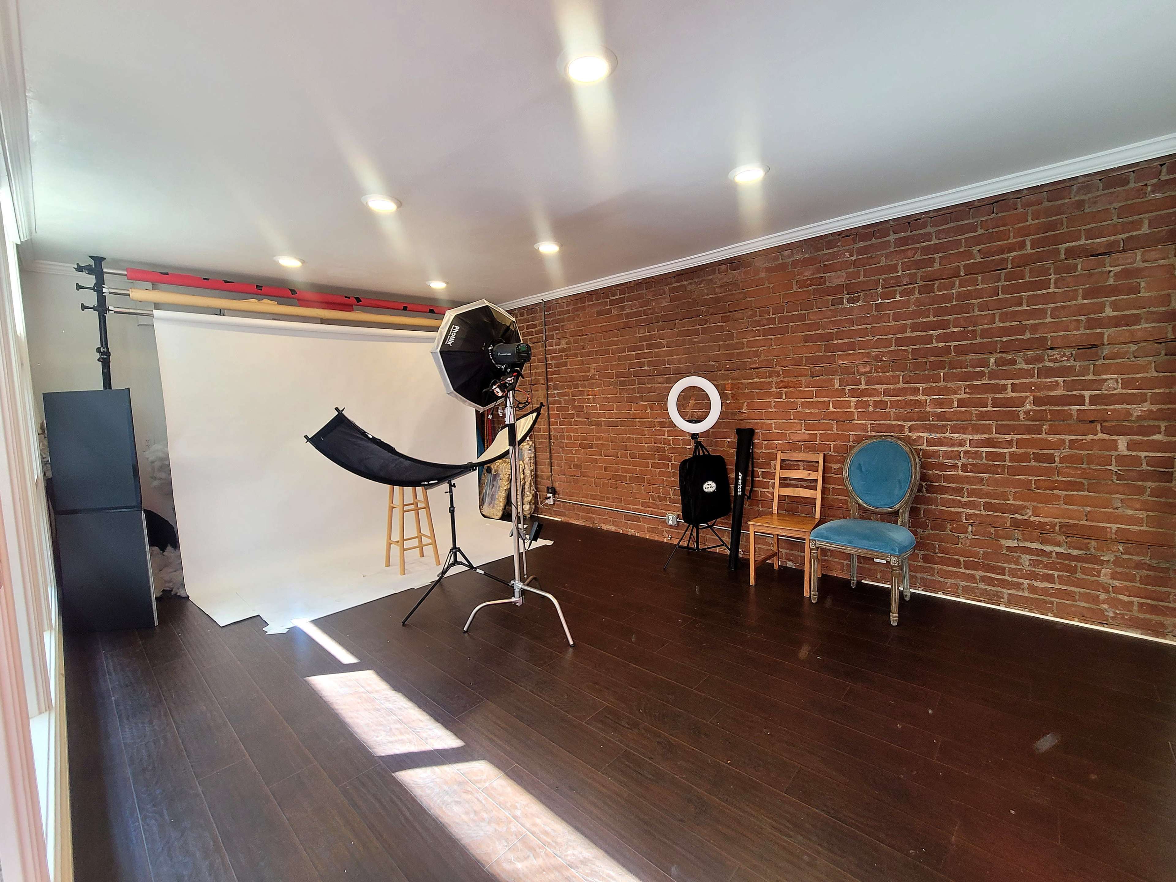 The image shows a photography studio with a backdrop, lighting equipment, and two chairs in a room with exposed brick walls and wooden flooring.