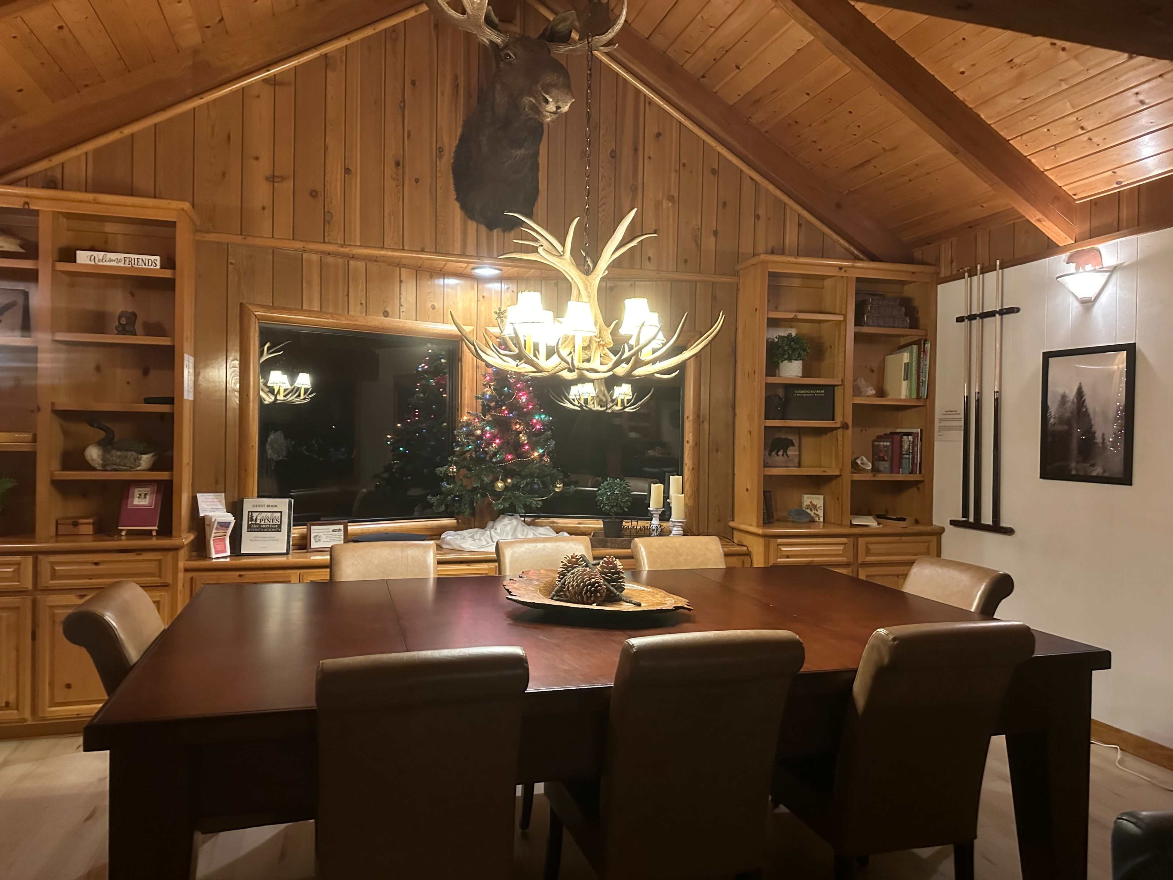 The image shows a warm, wooden dining room featuring a large table surrounded by chairs, with a chandelier above and a decorative moose head mounted on the wall.