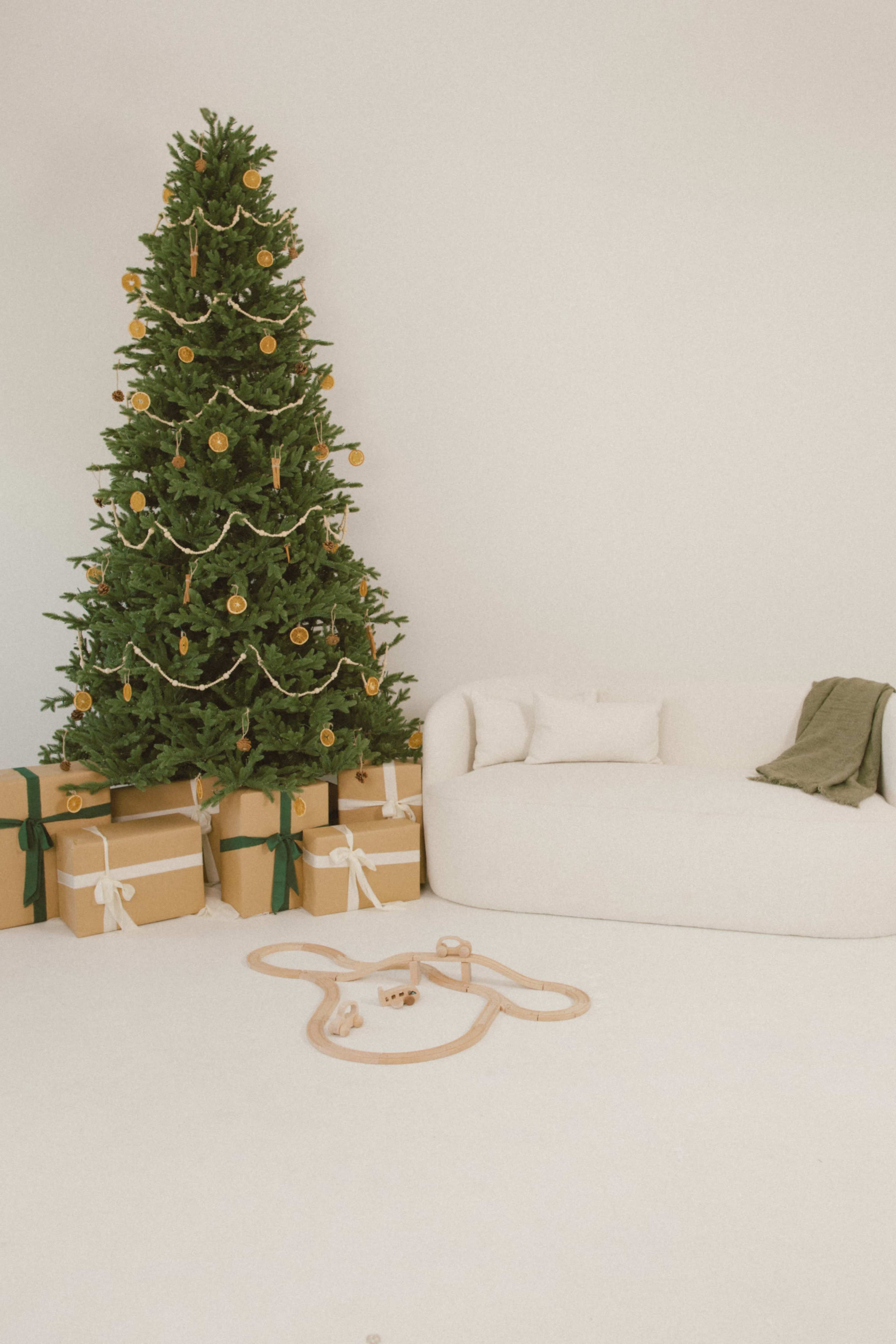 A Christmas tree decorated with ornaments and surrounded by wrapped gifts stands next to a light-colored couch and a wooden train set on the floor.