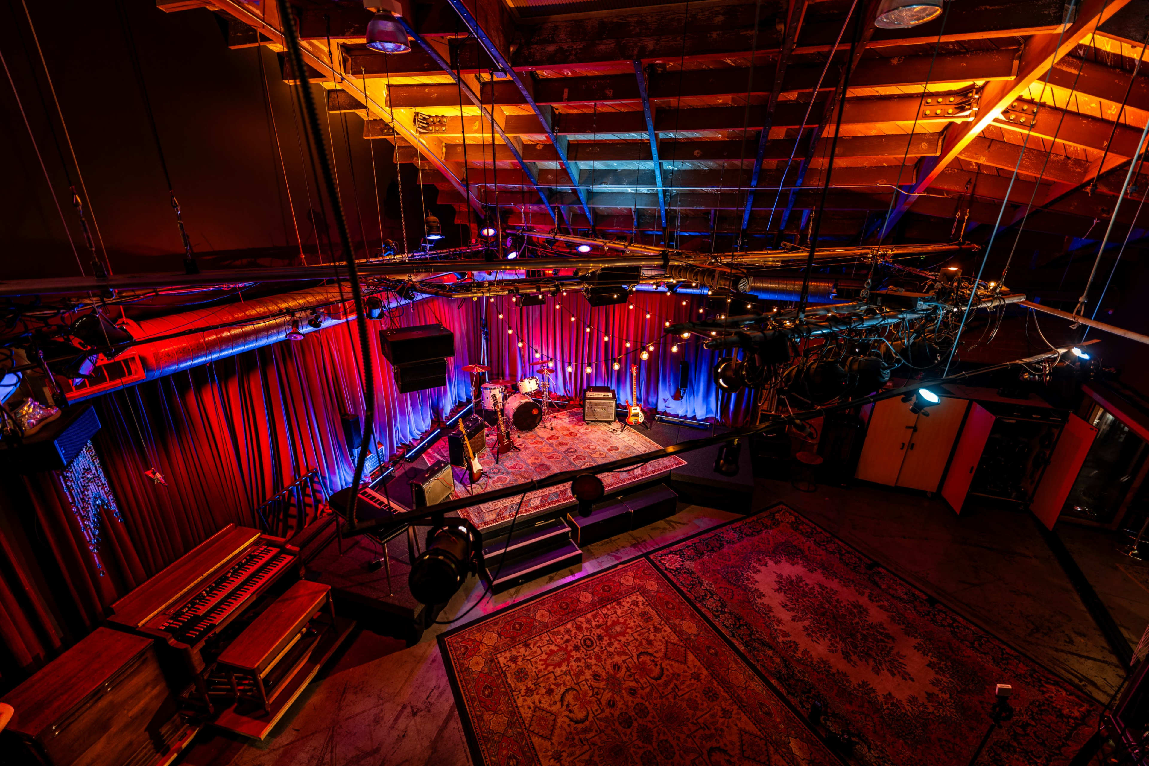 The image shows a brightly lit music performance space with colorful lighting, featuring a stage with instruments and a patterned rug.