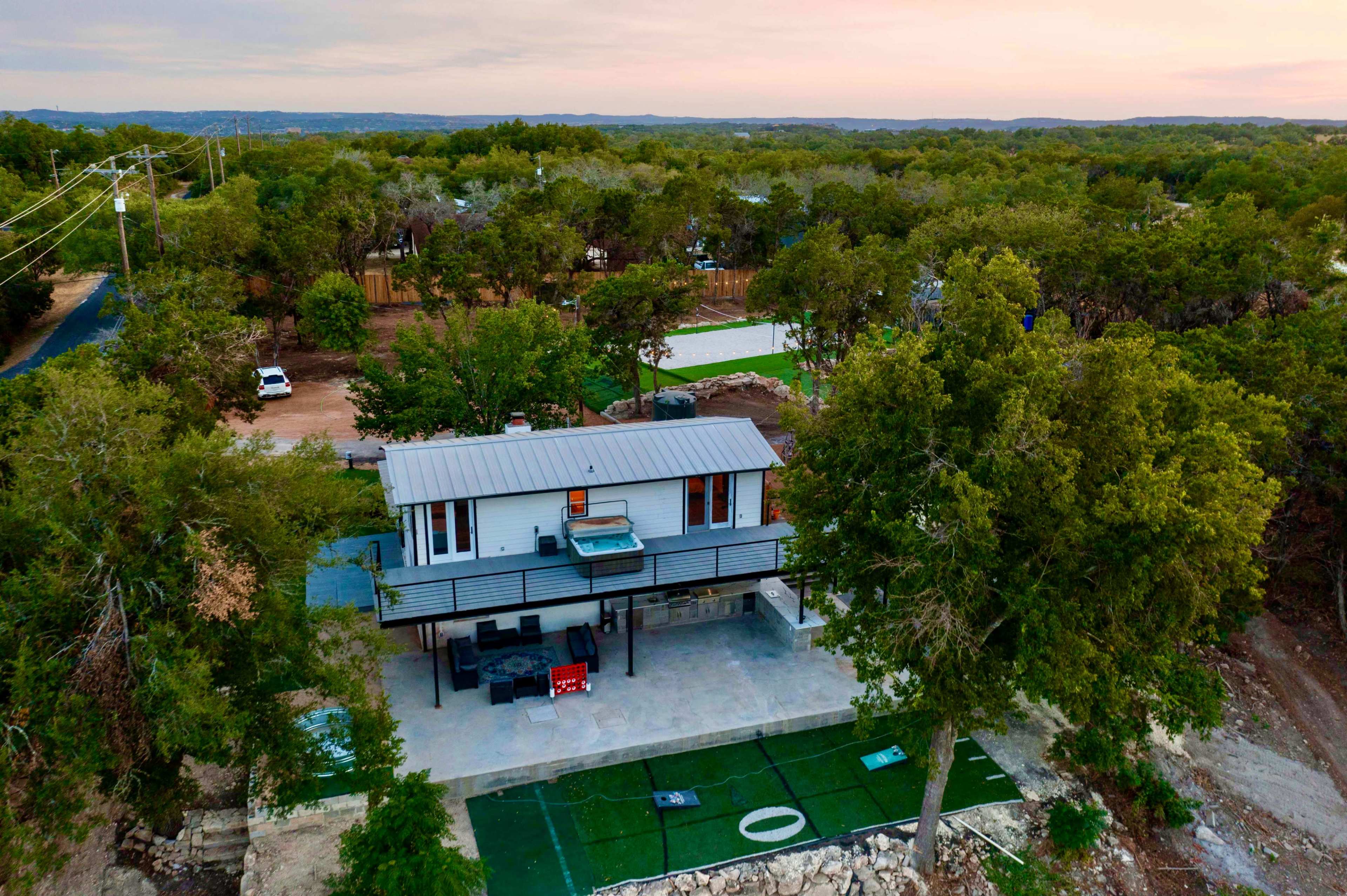 A modern house elevated on stilts, surrounded by trees and open land, with an outdoor terrace and putting green below.