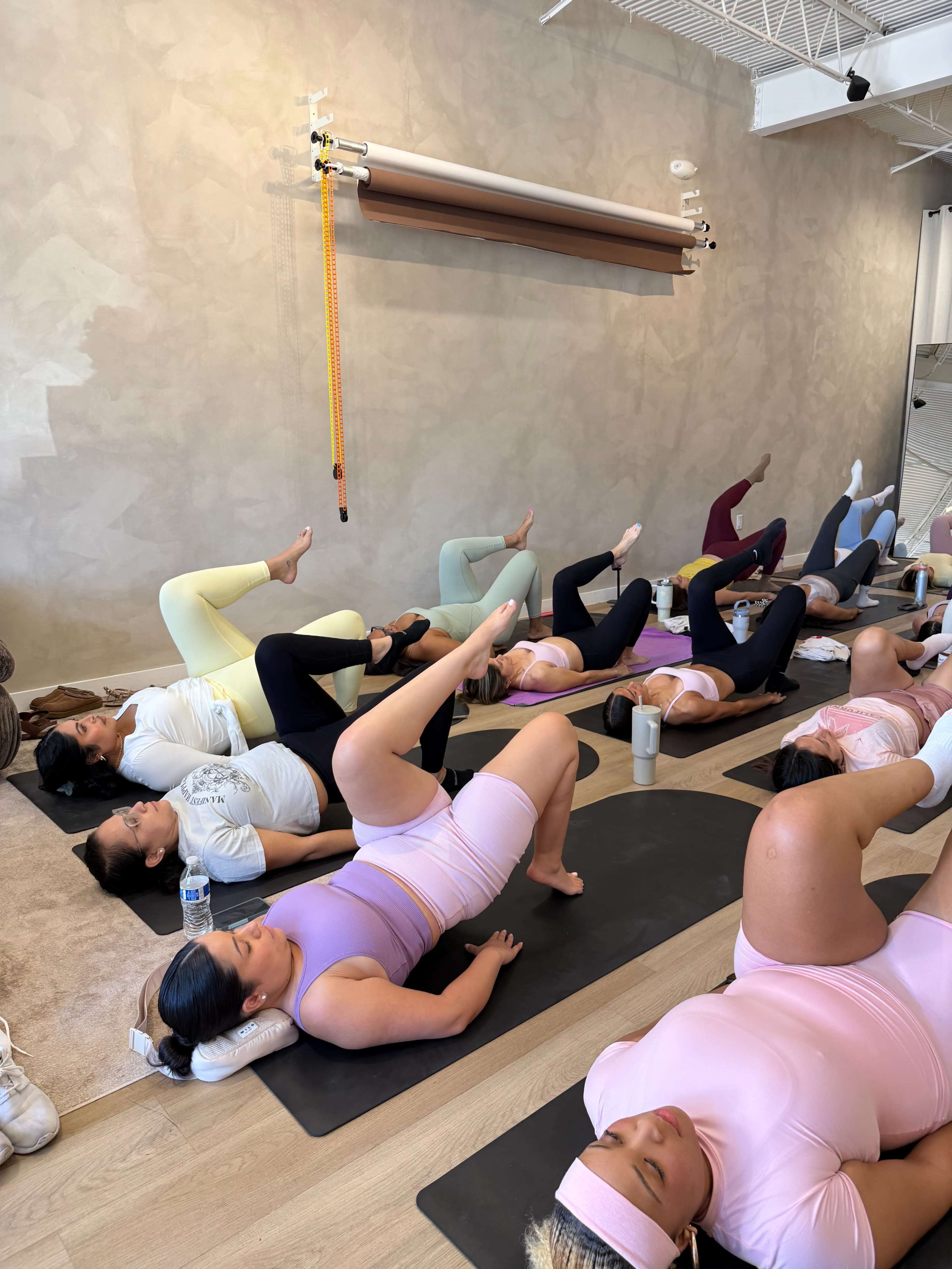 A group of individuals is participating in a yoga or fitness class on mats in a studio, performing exercises while lying on their backs with legs raised.