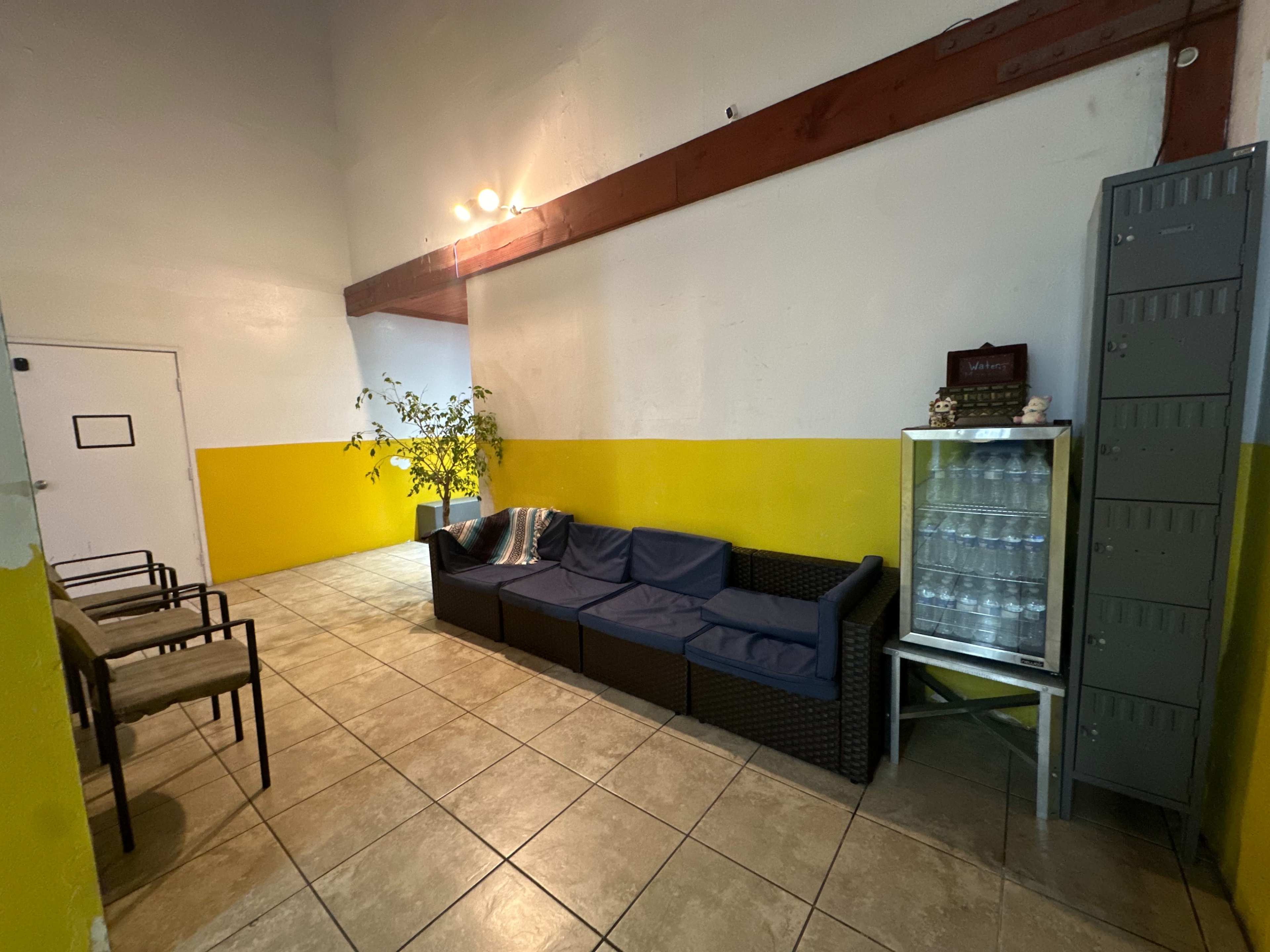 The image shows a hallway with yellow and white walls, featuring a seating area with a dark couch, chairs, a small plant, and a glass cabinet with bottles.