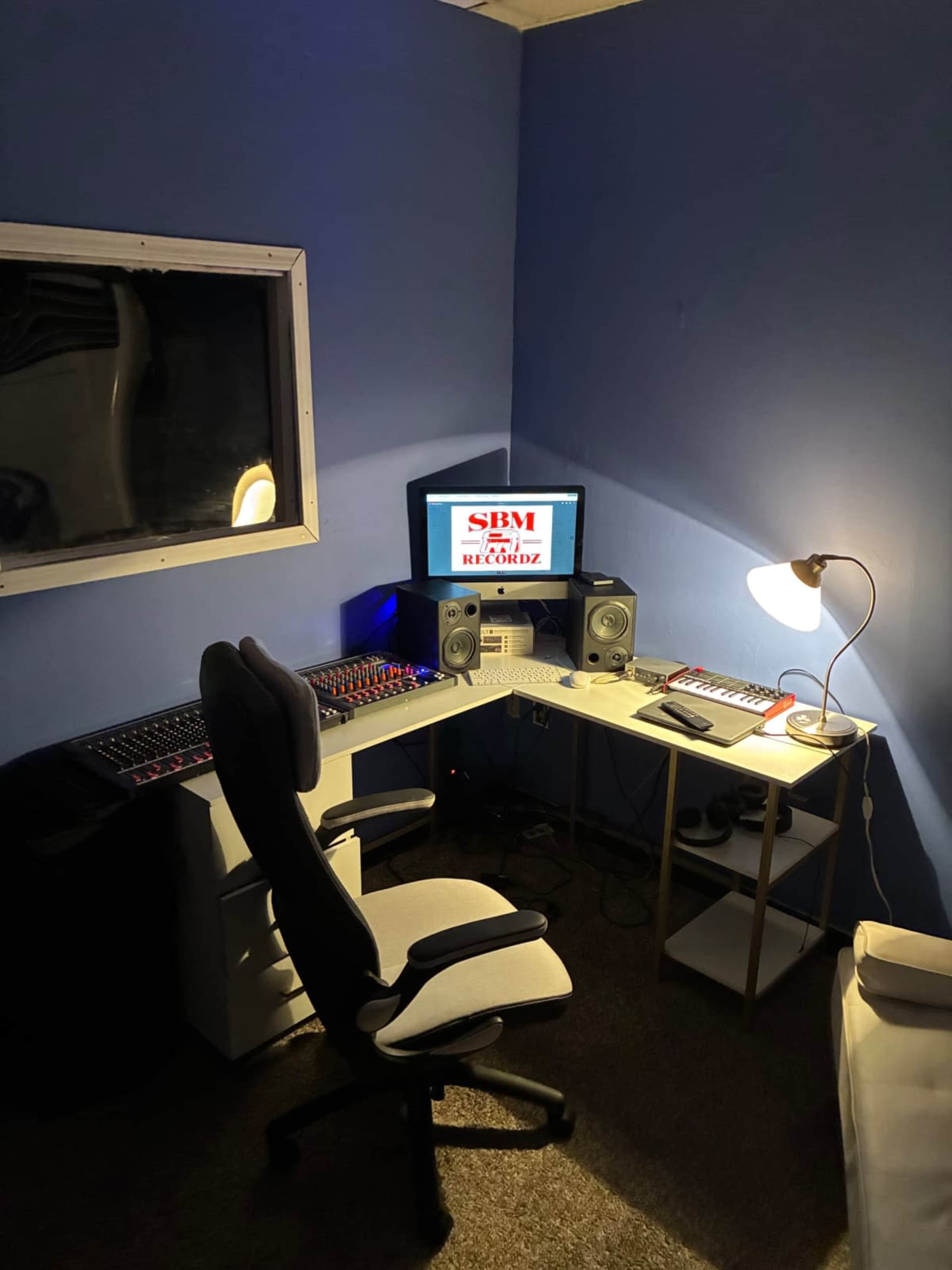 The image shows a small music recording studio featuring a desk with a computer, audio monitors, a keyboard, and a chair, all set against a blue wall.