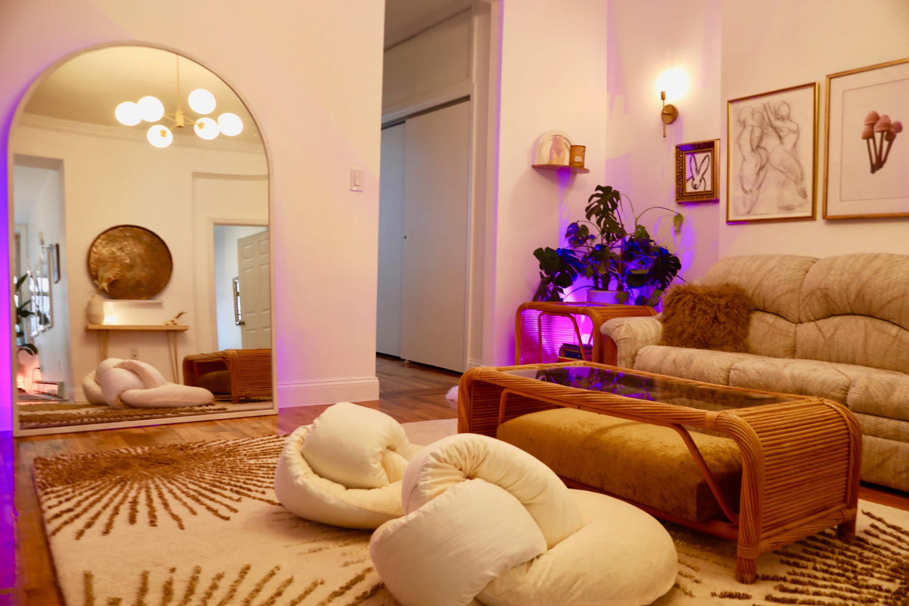 A cozy living room features a beige sofa, a glass coffee table atop a decorative rug, and a large mirror reflecting a well-lit space with indoor plants and purple accent lighting.