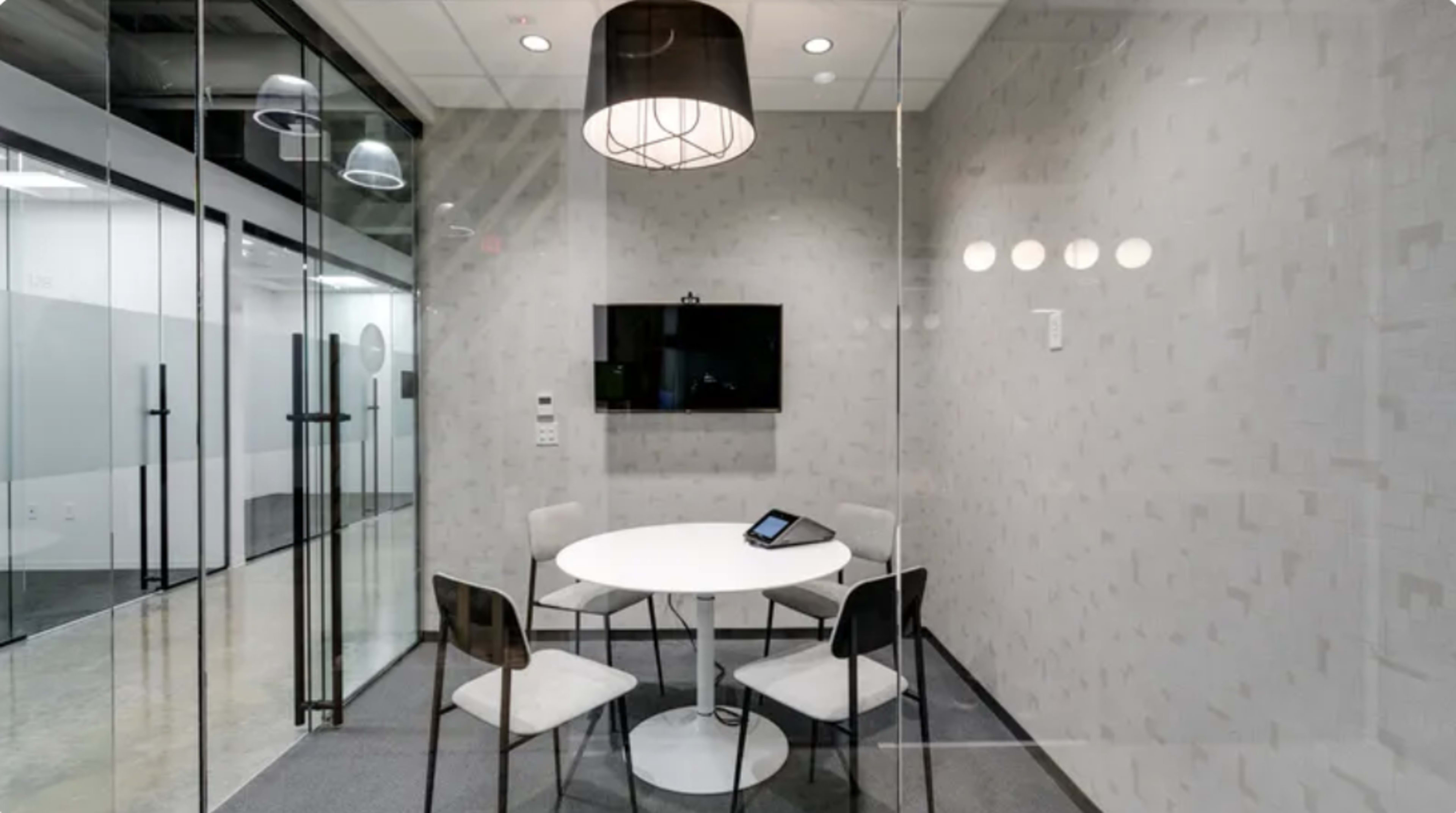 The image shows a small, enclosed meeting room featuring a round white table, four chairs, a wall-mounted screen, and large glass walls.