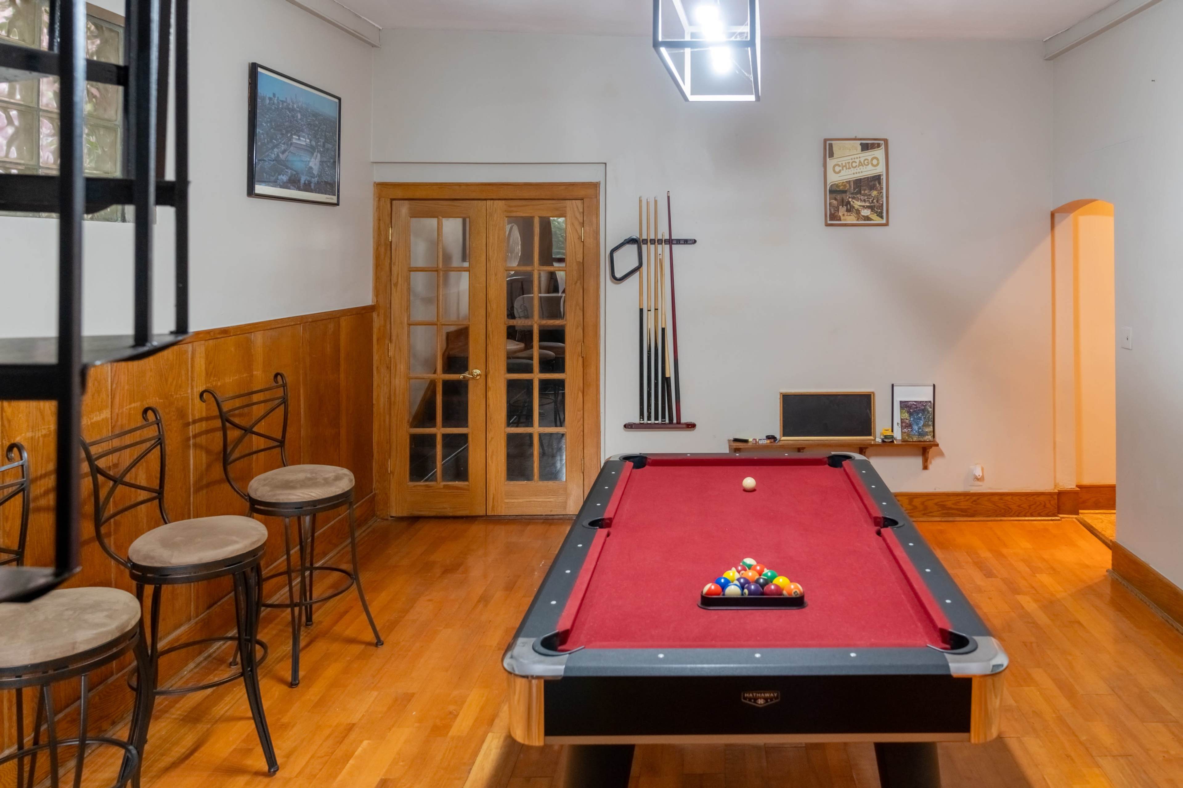 A pool room featuring a red felt billiard table, wooden flooring, and a set of bar stools against the wall.