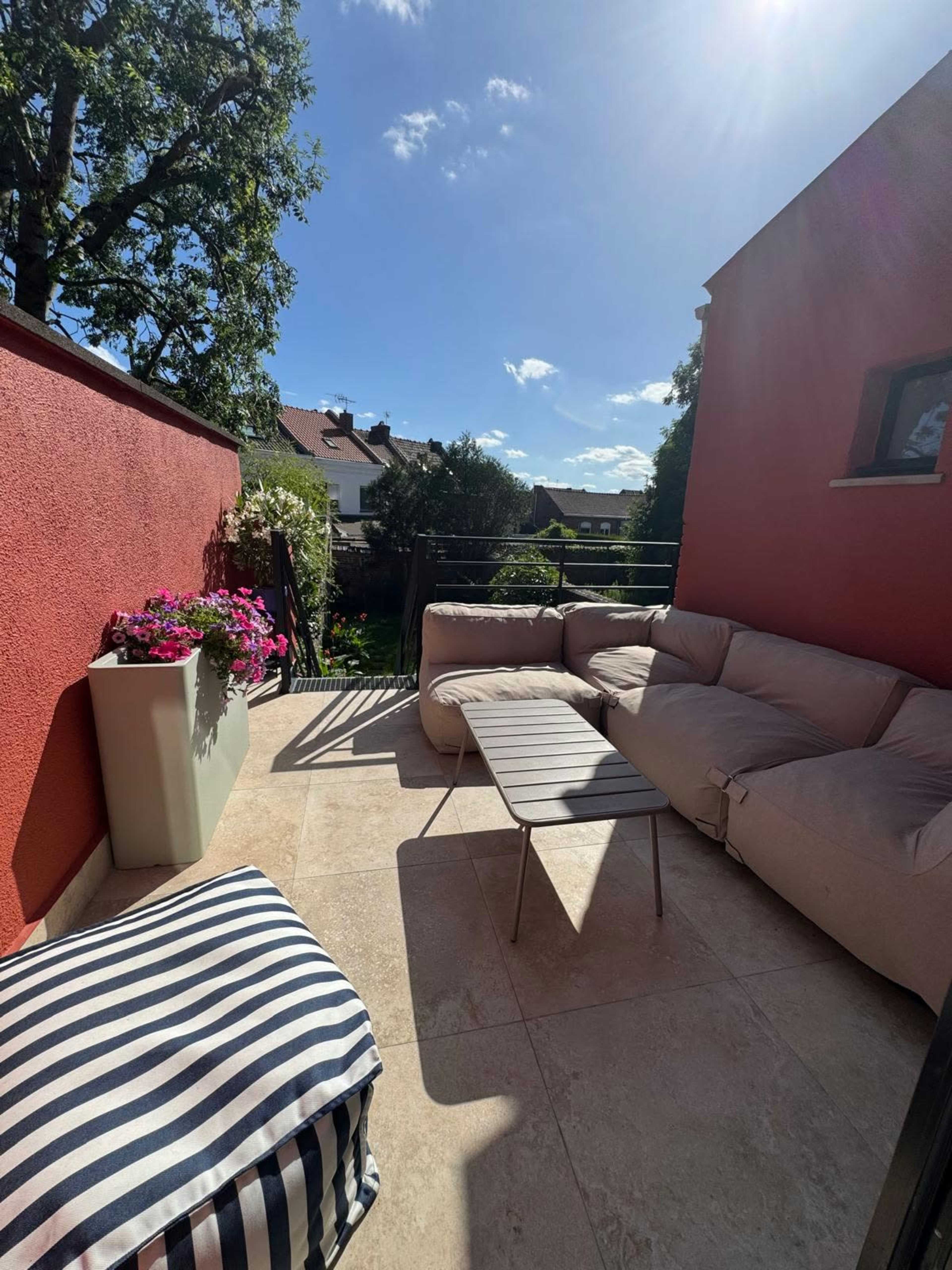 The image shows a sunlit terrace featuring a beige sectional sofa, a small table, and a striped pouf, with potted flowers and greenery in the background.
