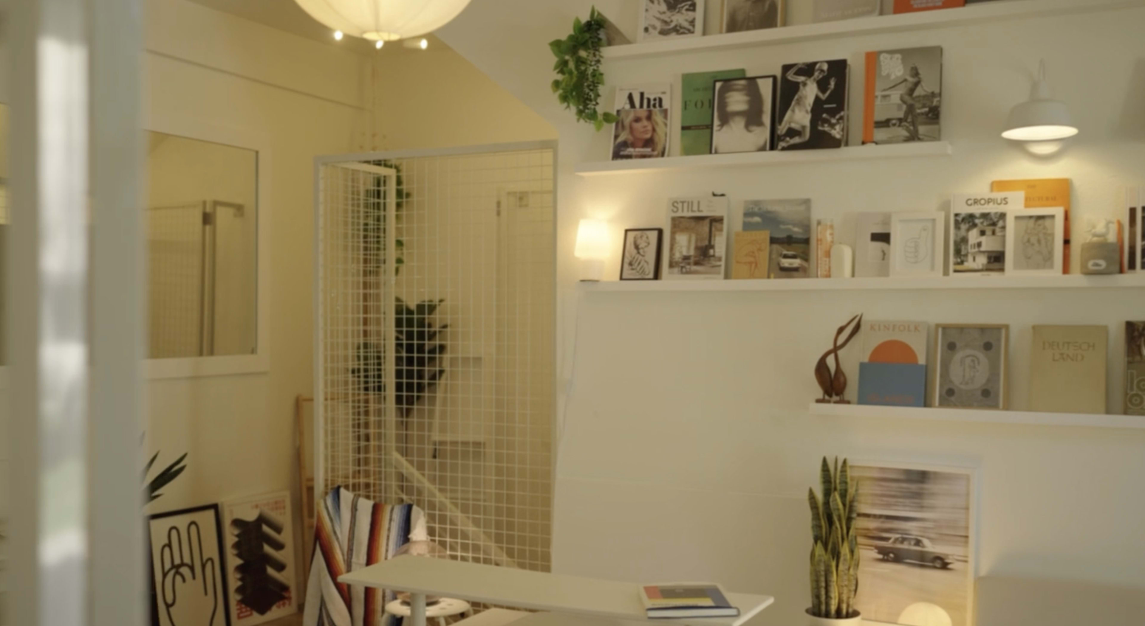 The image shows a cozy interior of a small shop or cafe, featuring a white wall with shelves displaying various books and magazines, a table in the center, and plants in decorative pots.