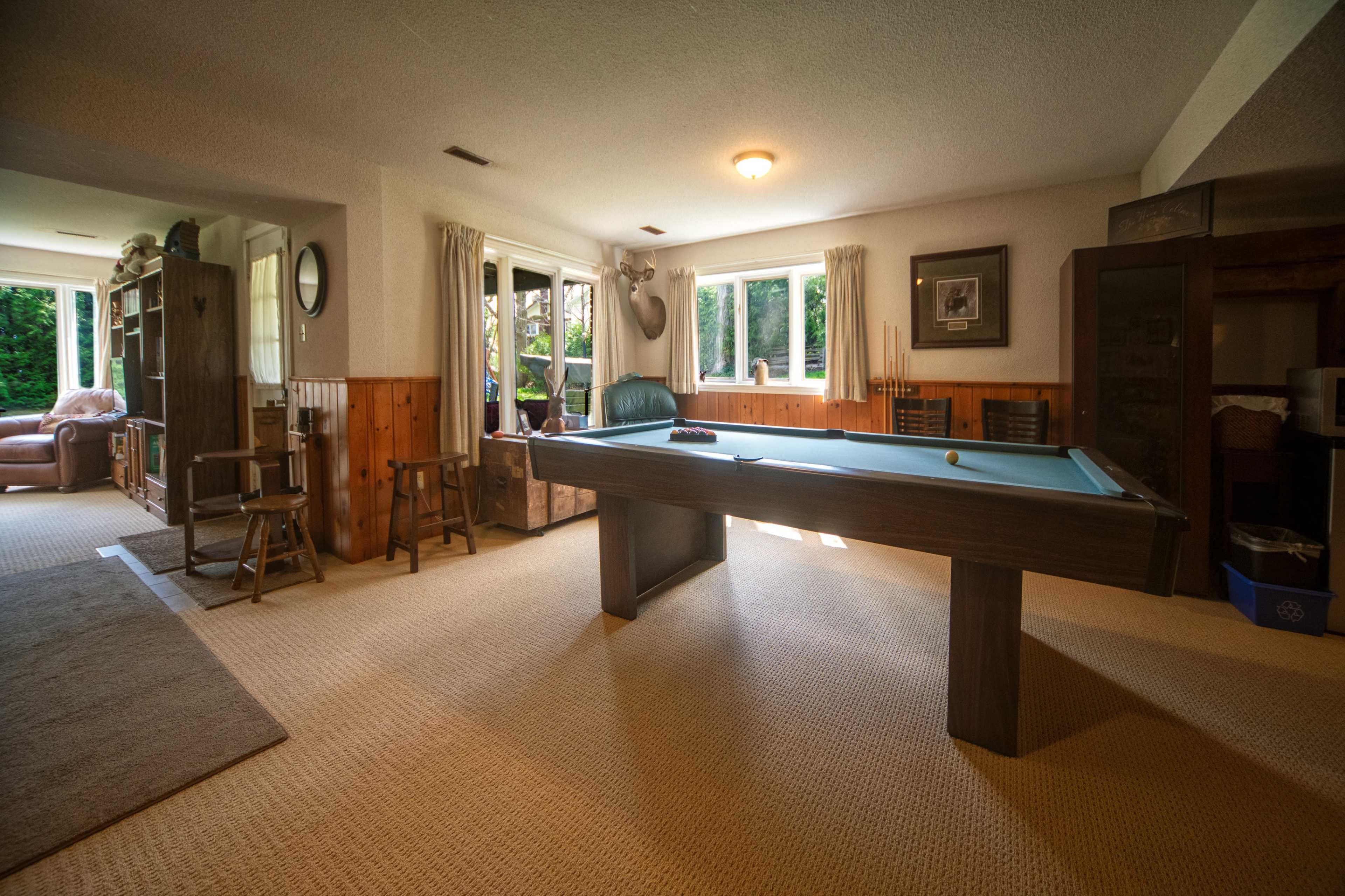 The image shows a room with a billiards table surrounded by a few chairs and a cozy seating area in the background, illuminated by natural light coming through large windows.