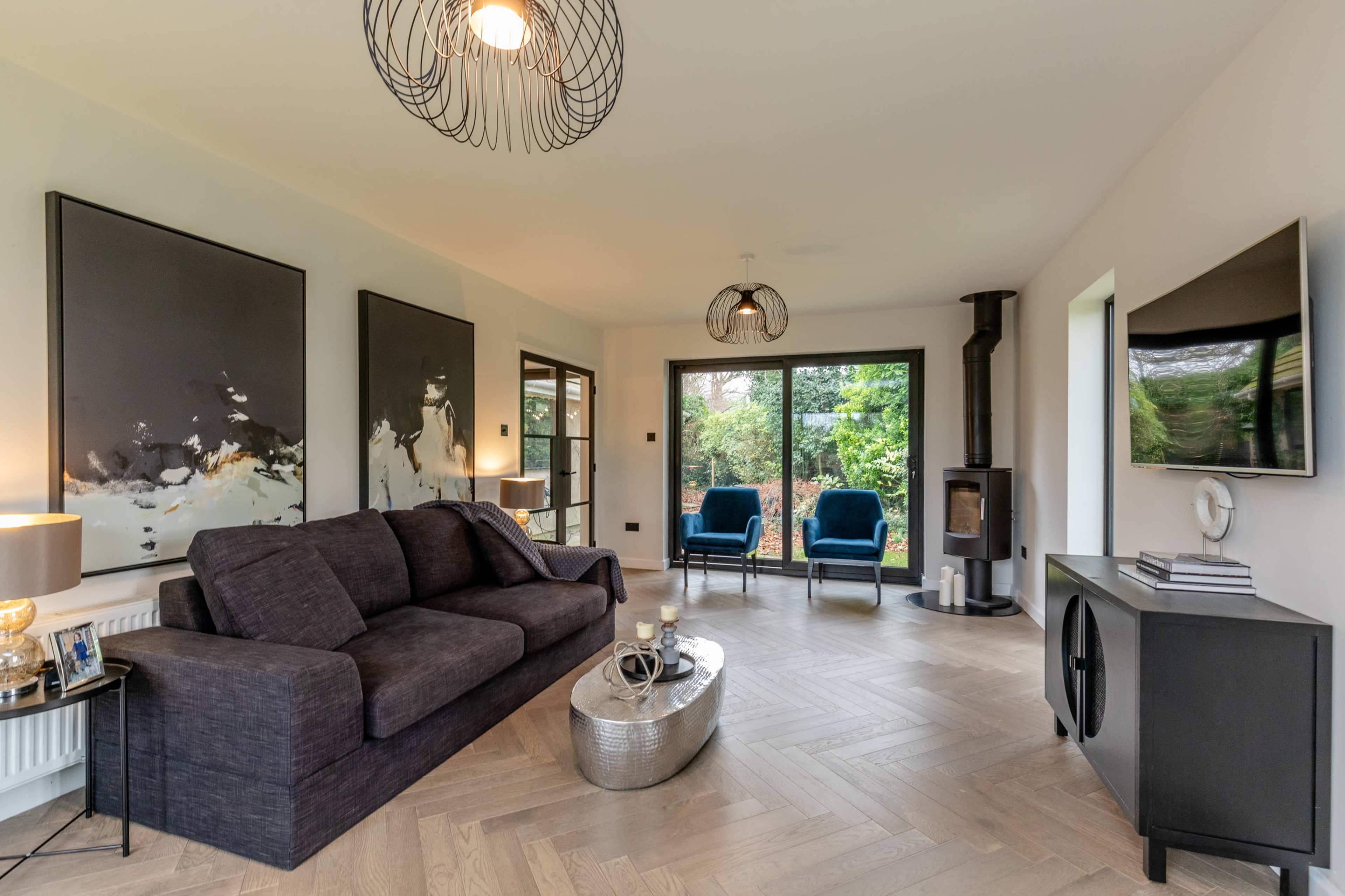 The image shows a modern living room with a dark sofa, a circular coffee table, a television mounted on the wall, and large windows overlooking a garden.