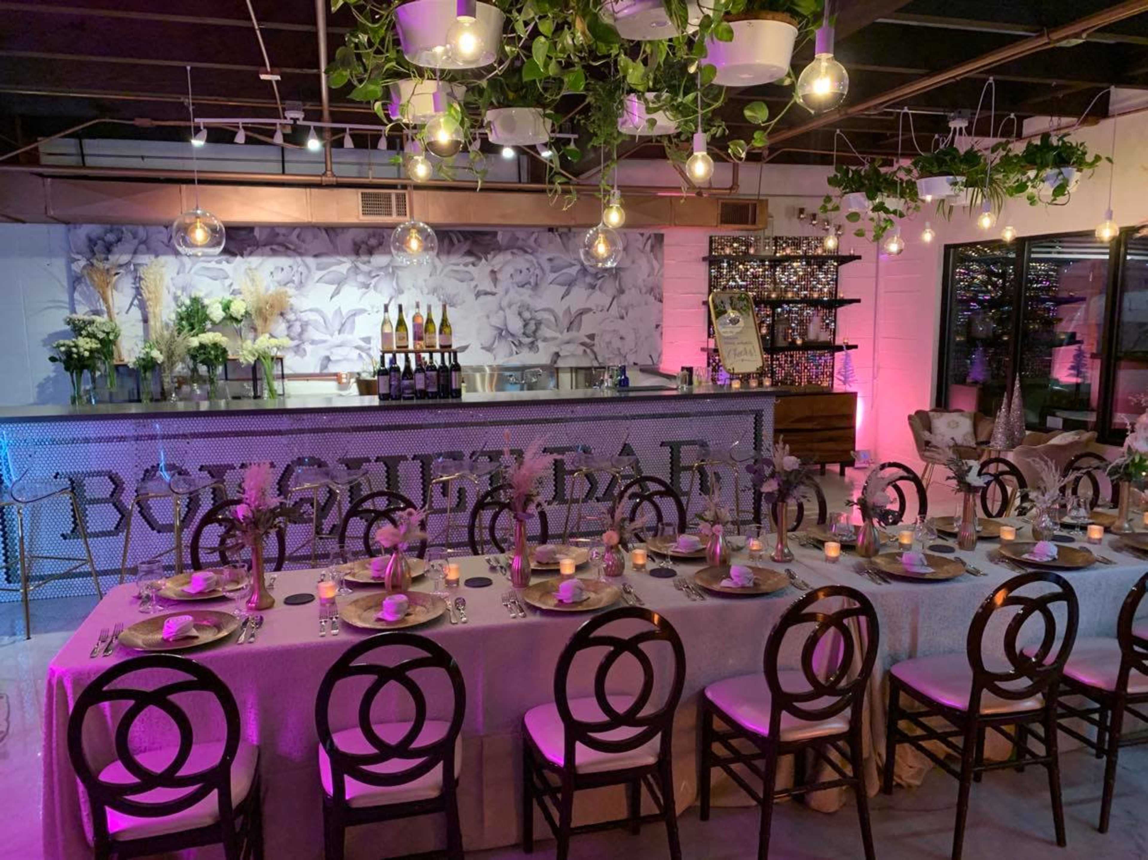 A long dining table is set with elegant tableware in a beautifully decorated bar area featuring a floral backdrop and illuminated with pink lights.