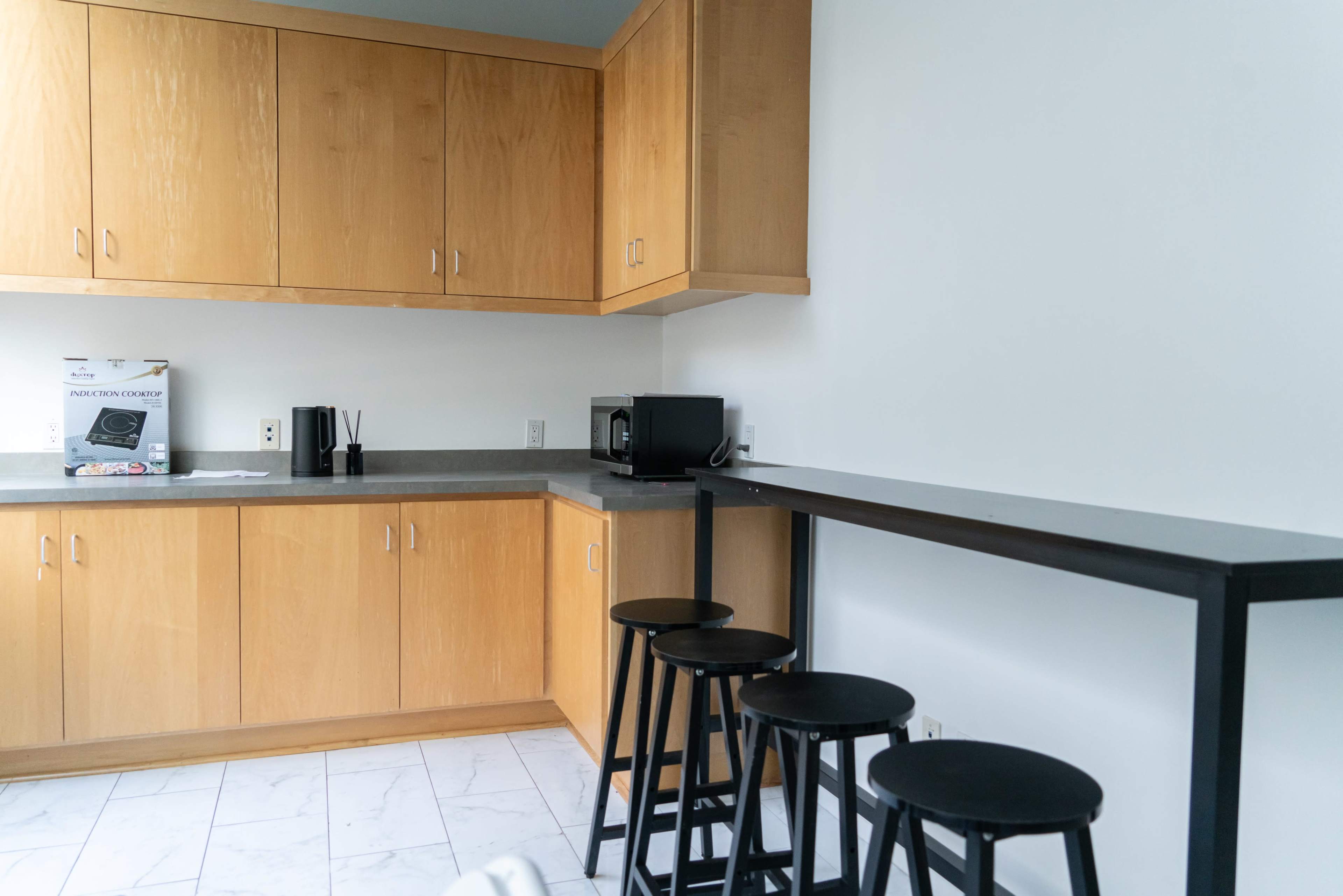 A small kitchen area features wooden cabinets, a gray countertop, a microwave, and four black bar stools beside a tall table.