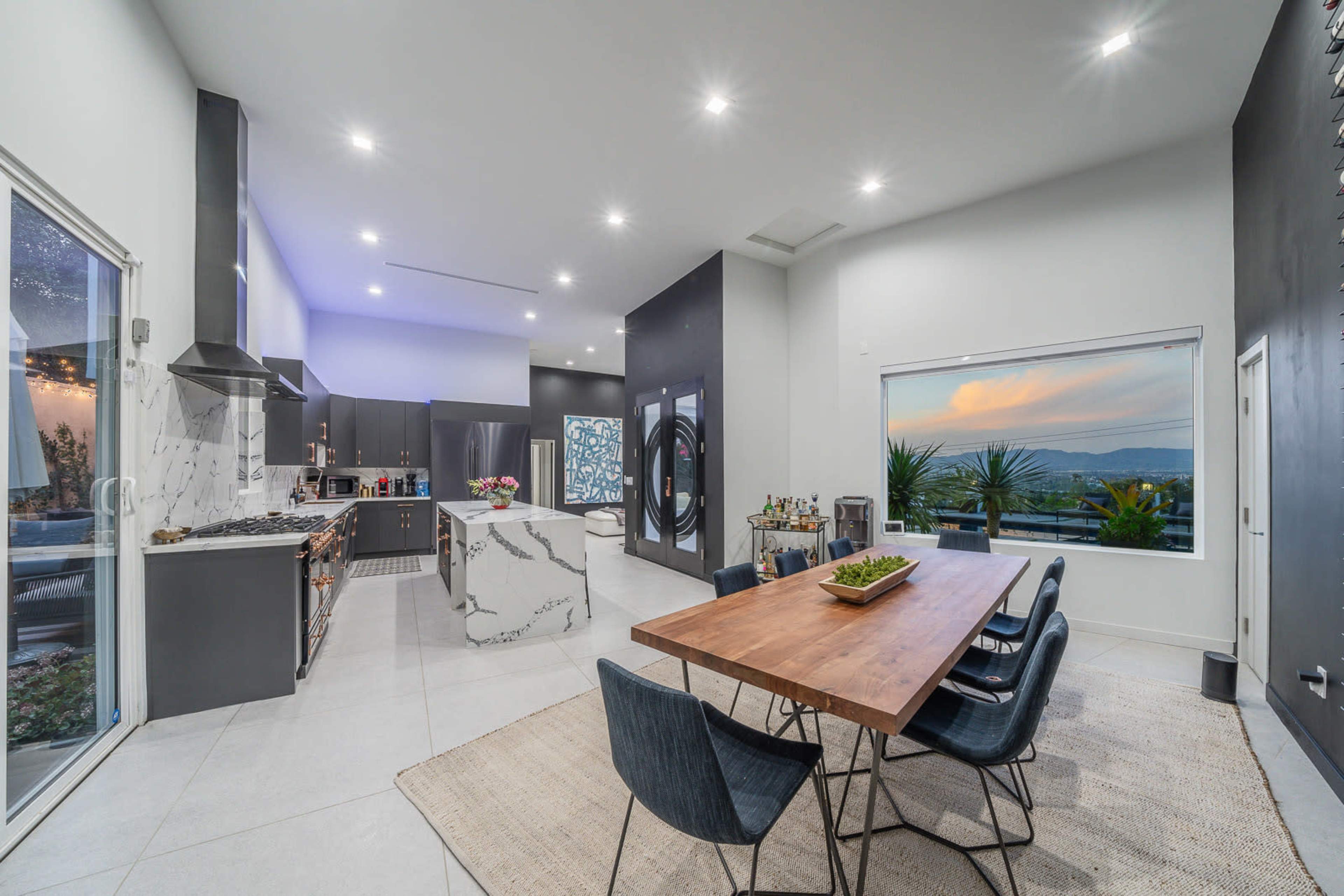 A modern kitchen and dining area feature a marble-topped table, sleek cabinetry, and large windows overlooking a sunset view.