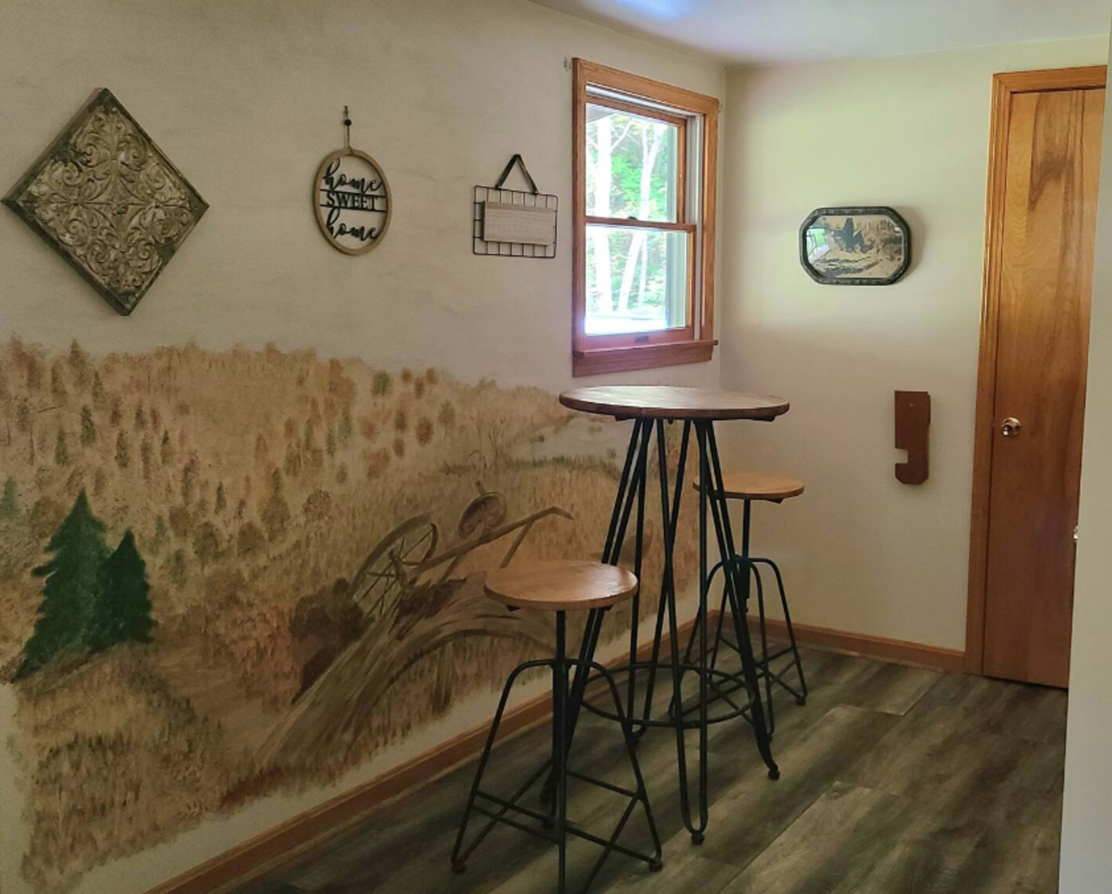 A small room featuring a wooden bar table with three stools and a wall mural depicting a landscape scene.