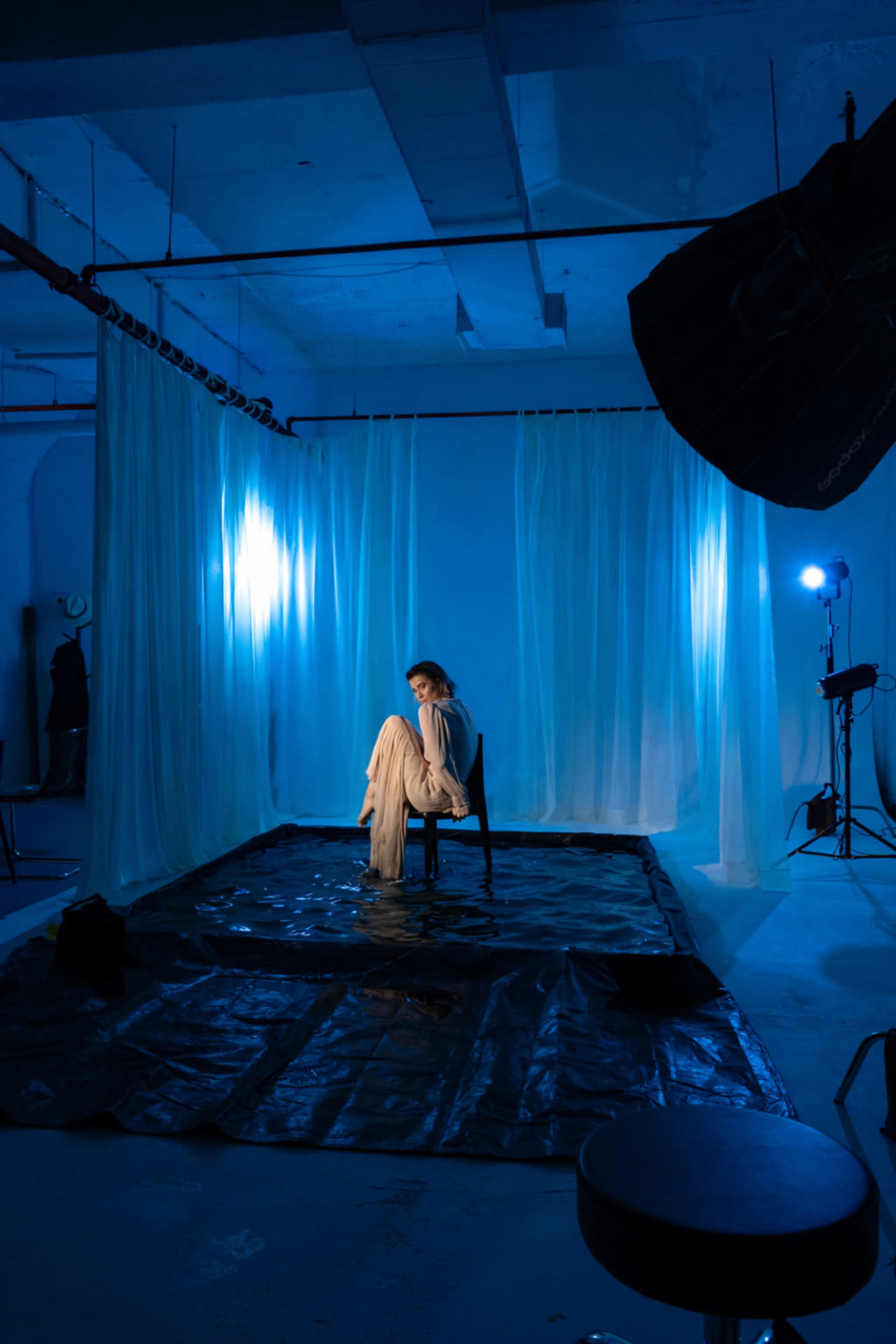 A model sits on a chair in a partially flooded studio surrounded by blue lighting and draped fabric.