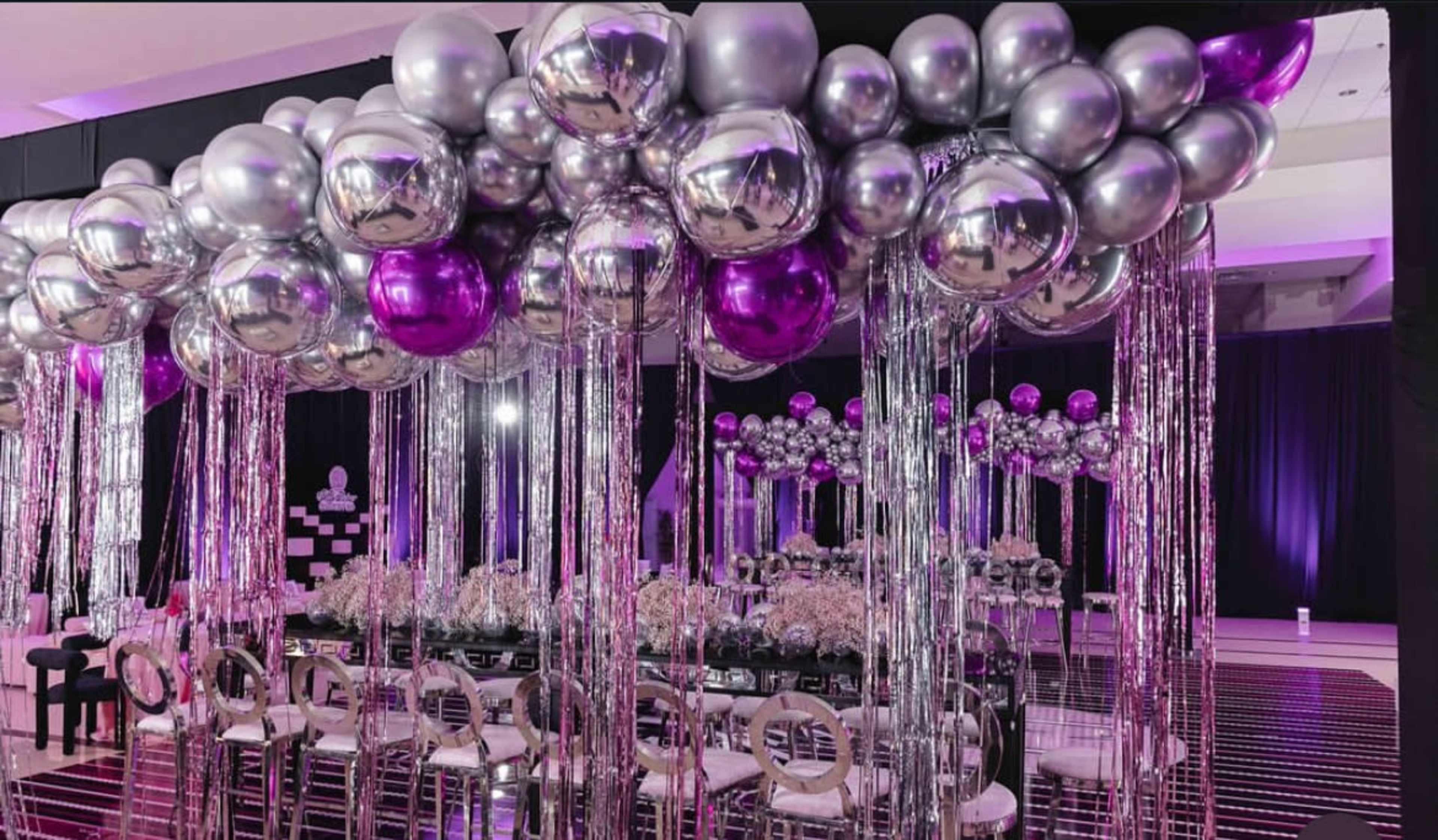 The image shows a decorative event space adorned with silver and purple balloons, along with shimmering fringe and elegant seating arrangements.