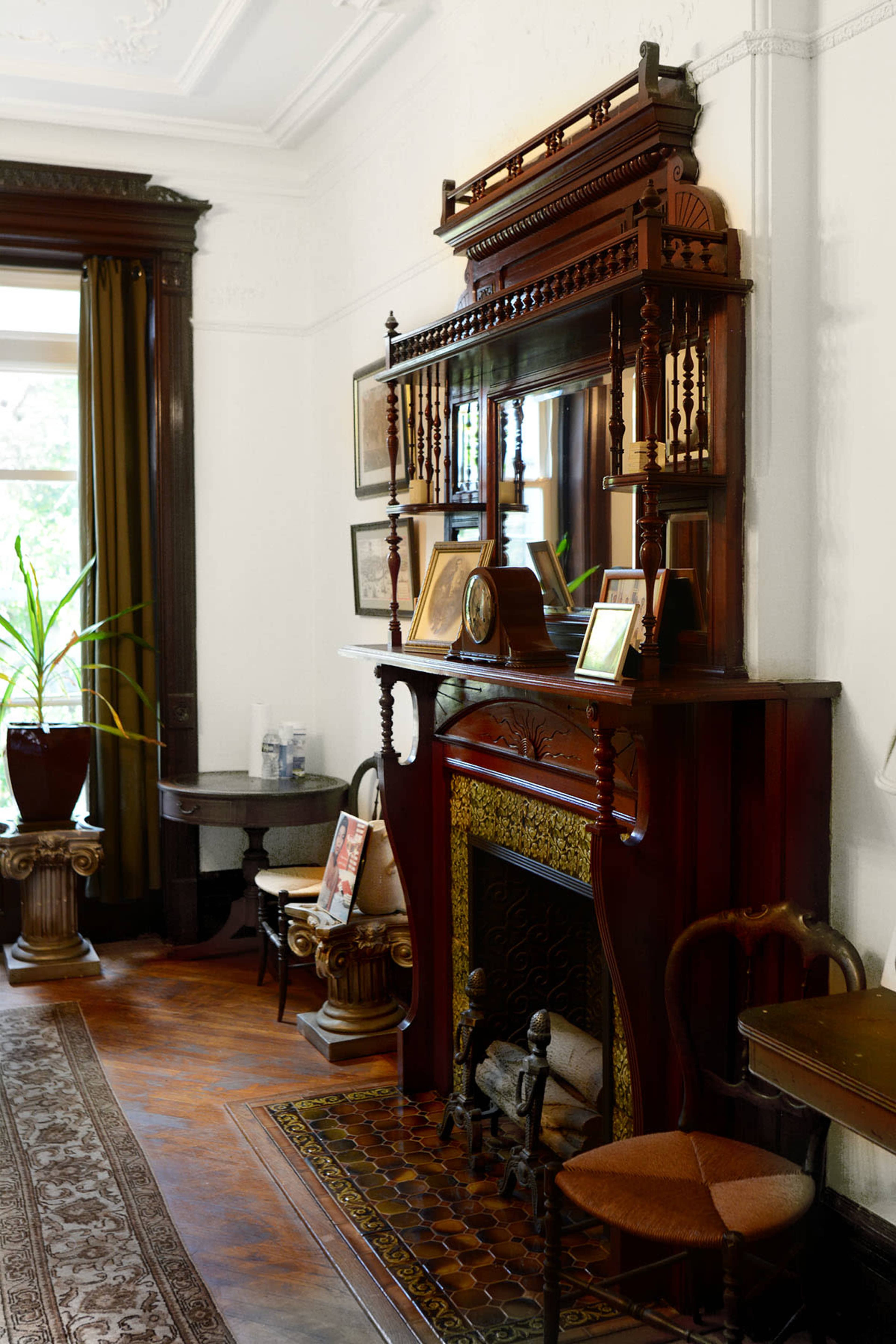 The image shows a vintage wooden fireplace with an ornate mantelpiece, surrounded by decorative furniture and plants in a well-lit room.