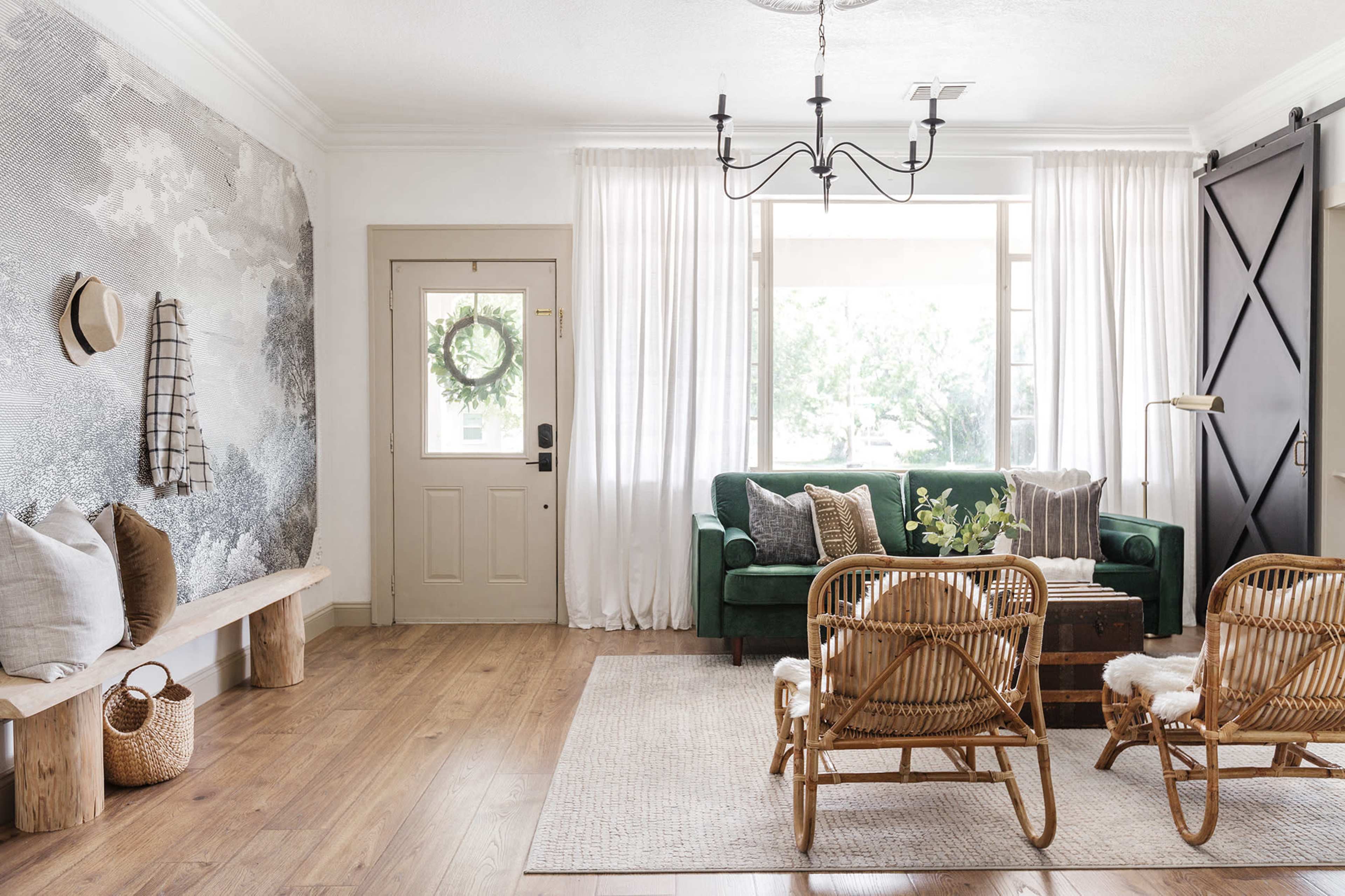 A bright living room features a green sofa, two rattan chairs, a wooden bench, and large windows with sheer curtains.