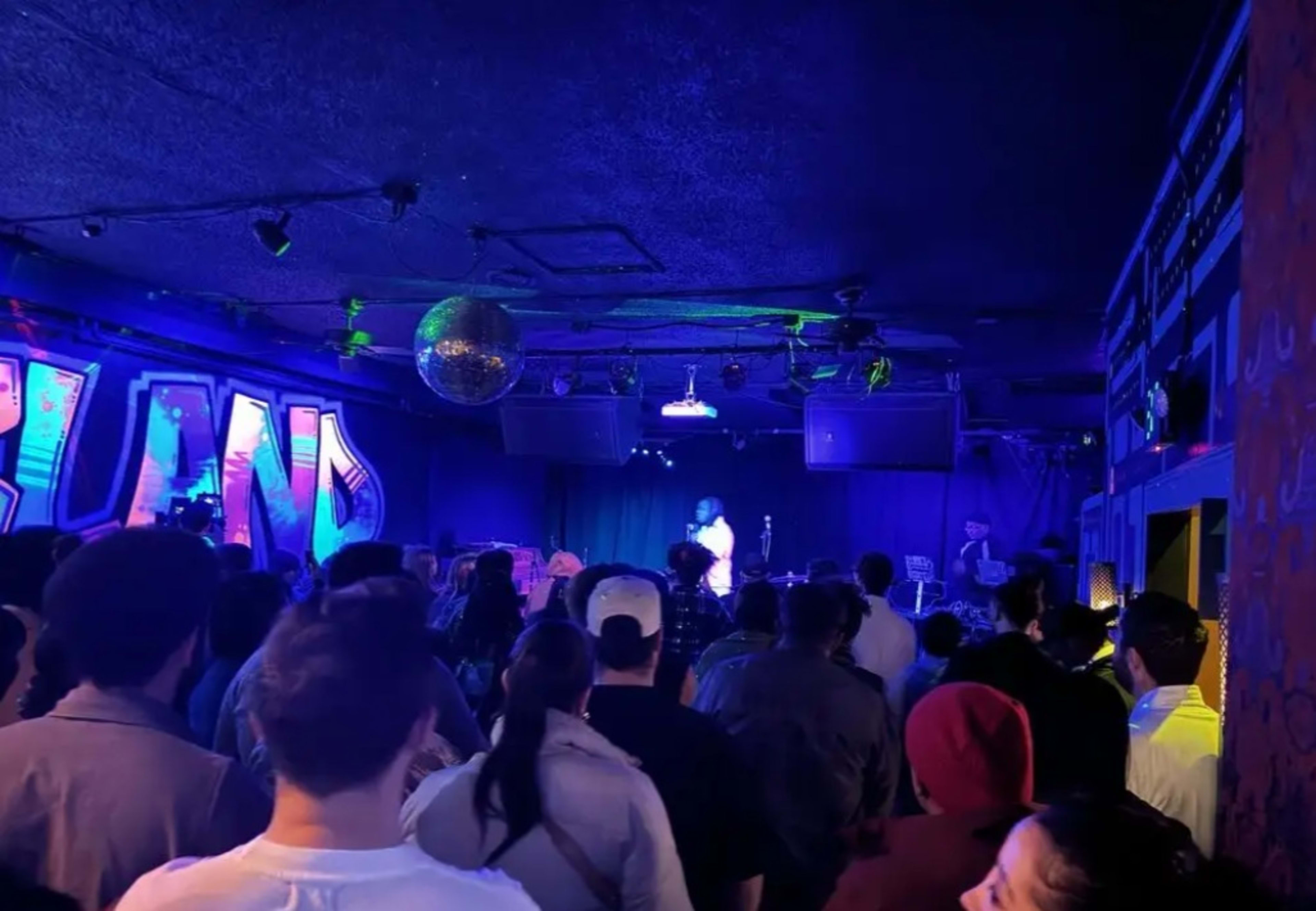 A crowd stands facing a performer on stage in a dimly lit venue adorned with colorful decorations and a disco ball.