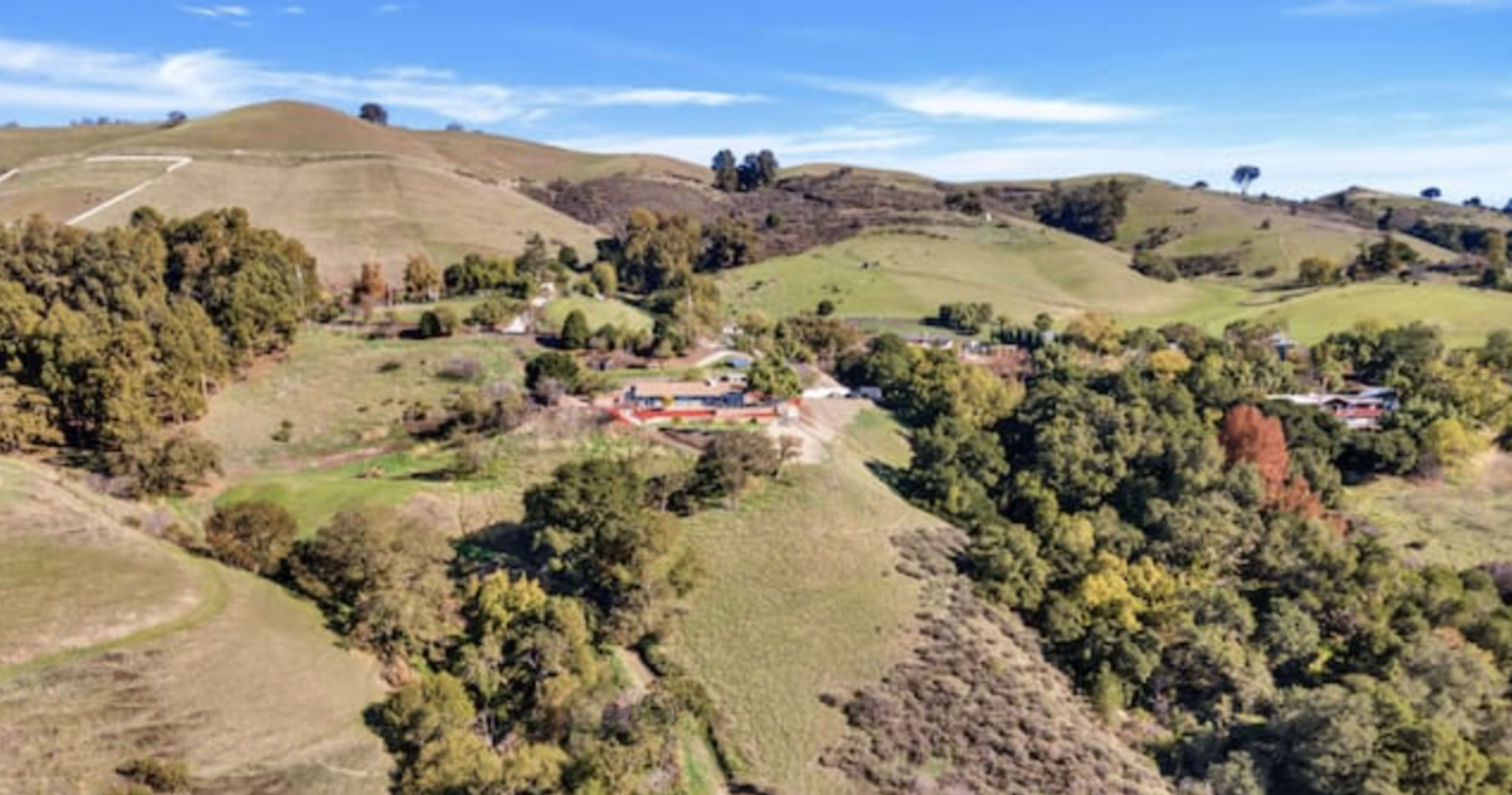 A rural landscape with rolling green hills, scattered trees, and a cluster of residential buildings in the foreground.