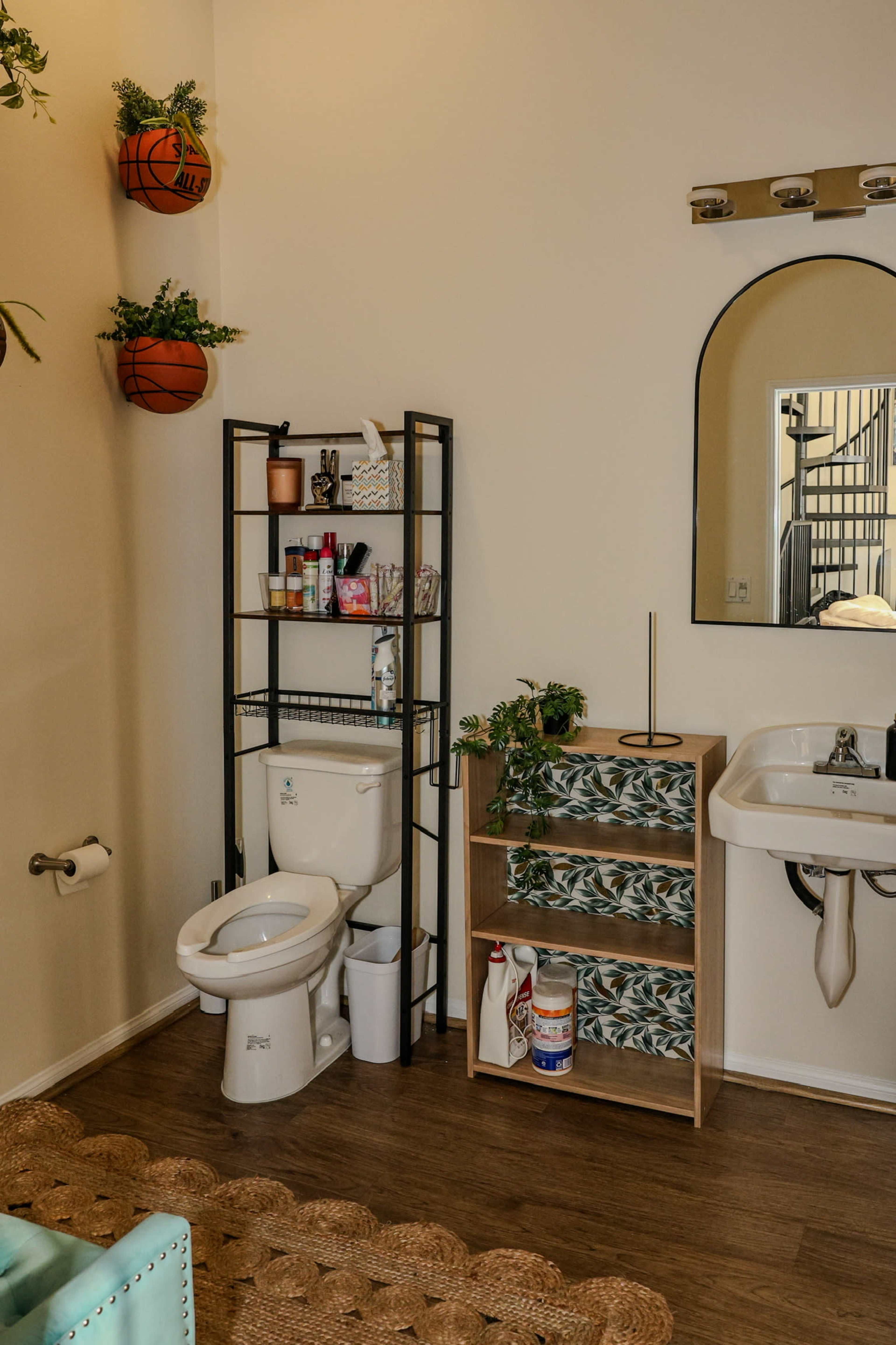 The image shows a bathroom area with a toilet, a small storage rack with personal care products, and a sink mounted on the wall, alongside a staircase in the background.