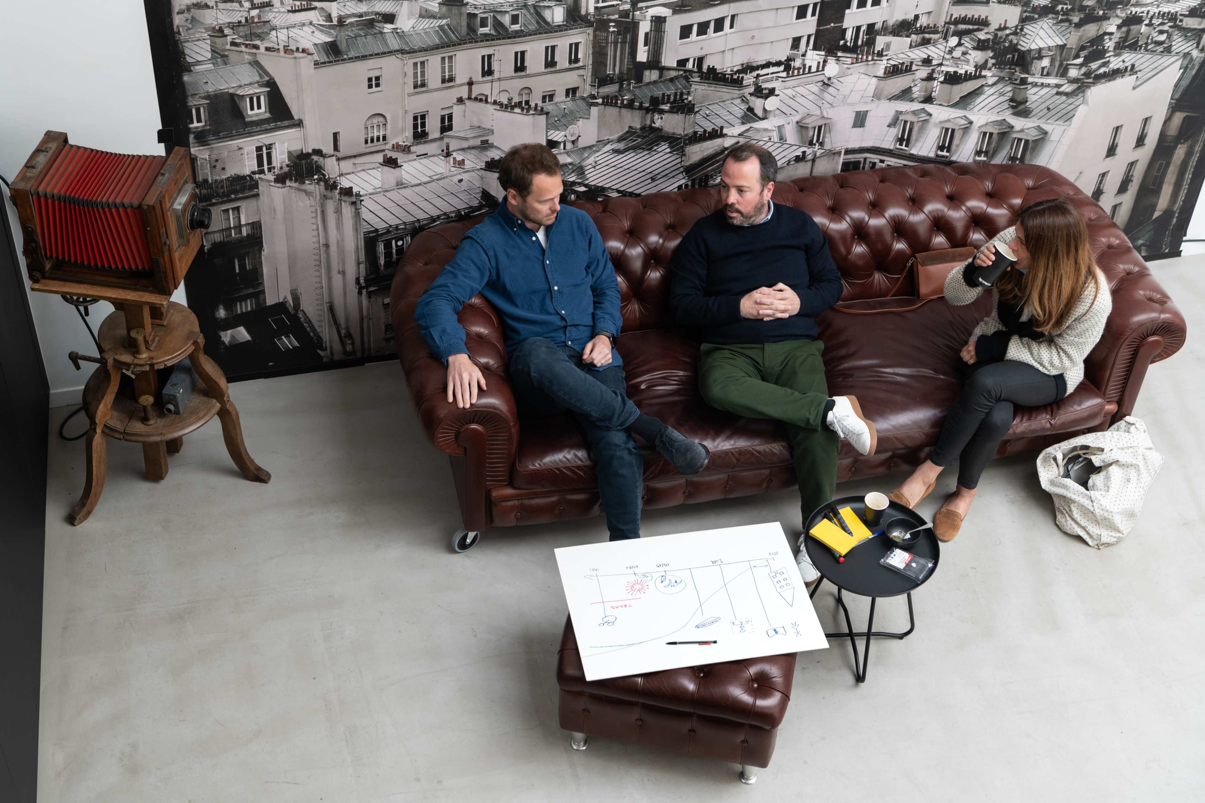 Three people sit on a brown leather couch in a modern workspace, discussing a project with a large sheet of paper and various tools placed on the floor.