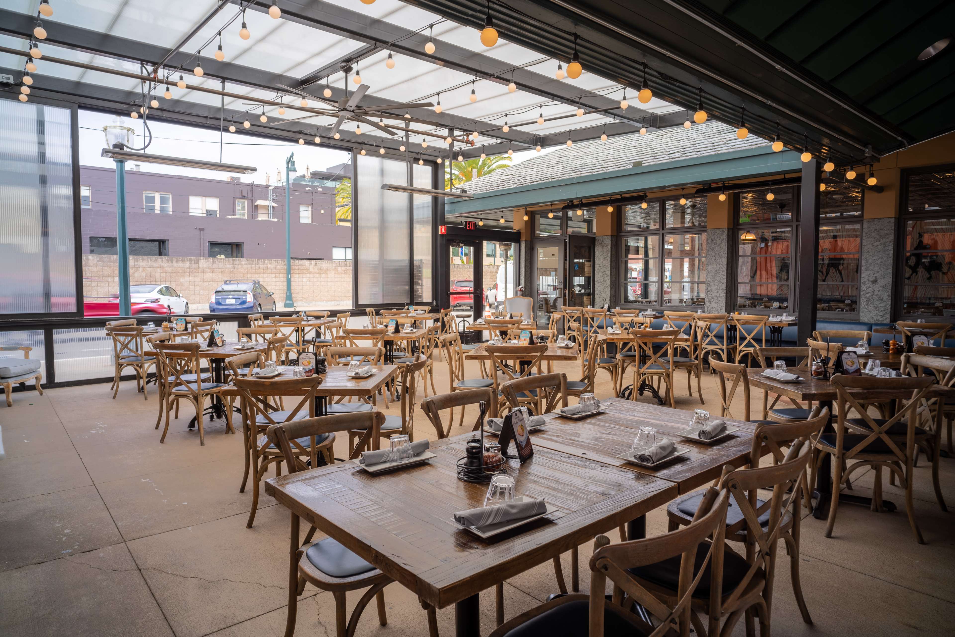 The image shows a spacious restaurant interior with wooden tables and chairs, arranged neatly for dining, and large windows allowing natural light.