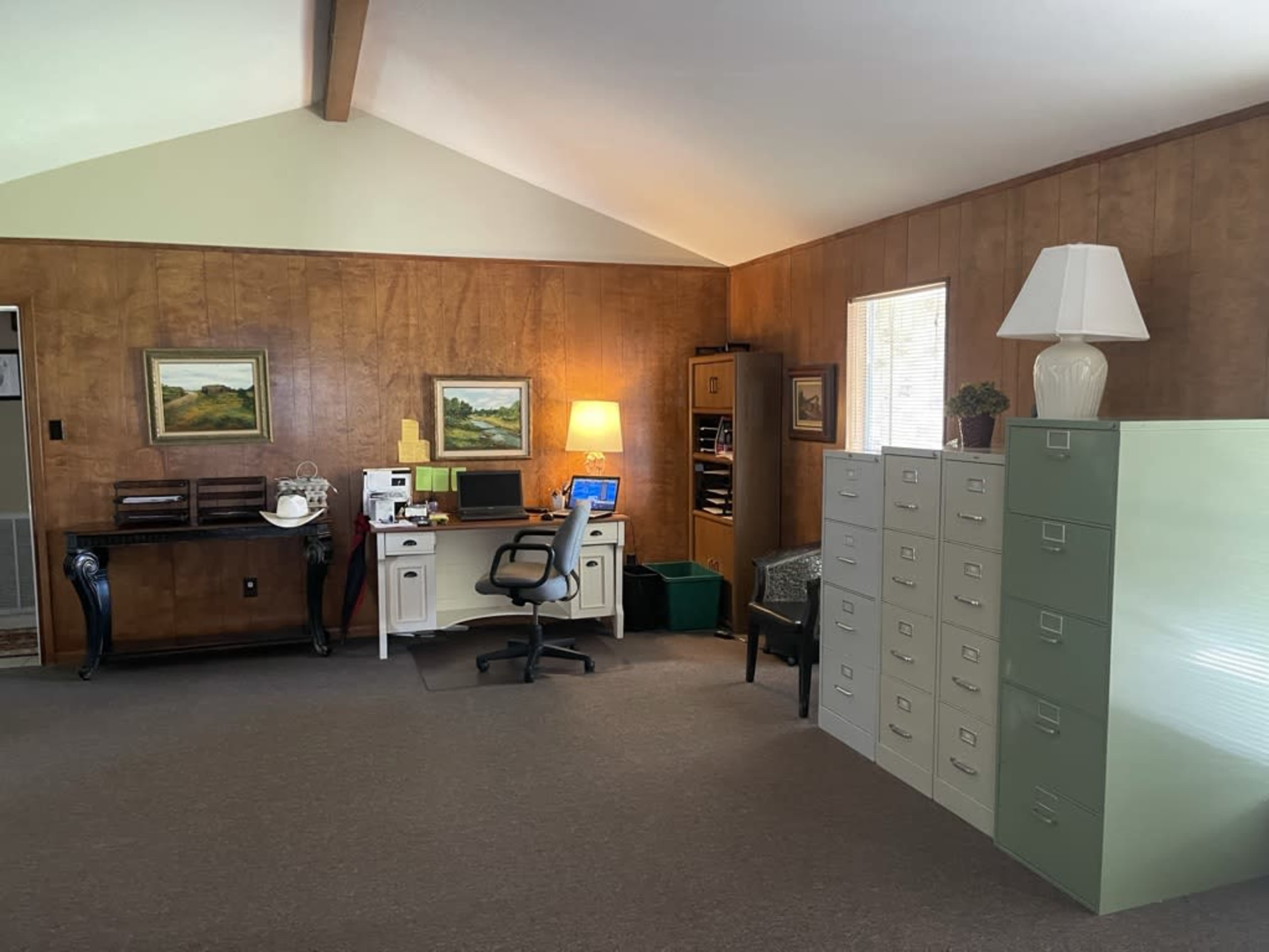 An office space with wooden walls, a desk with a computer and chair, filing cabinets, and soft lighting from a lamp.