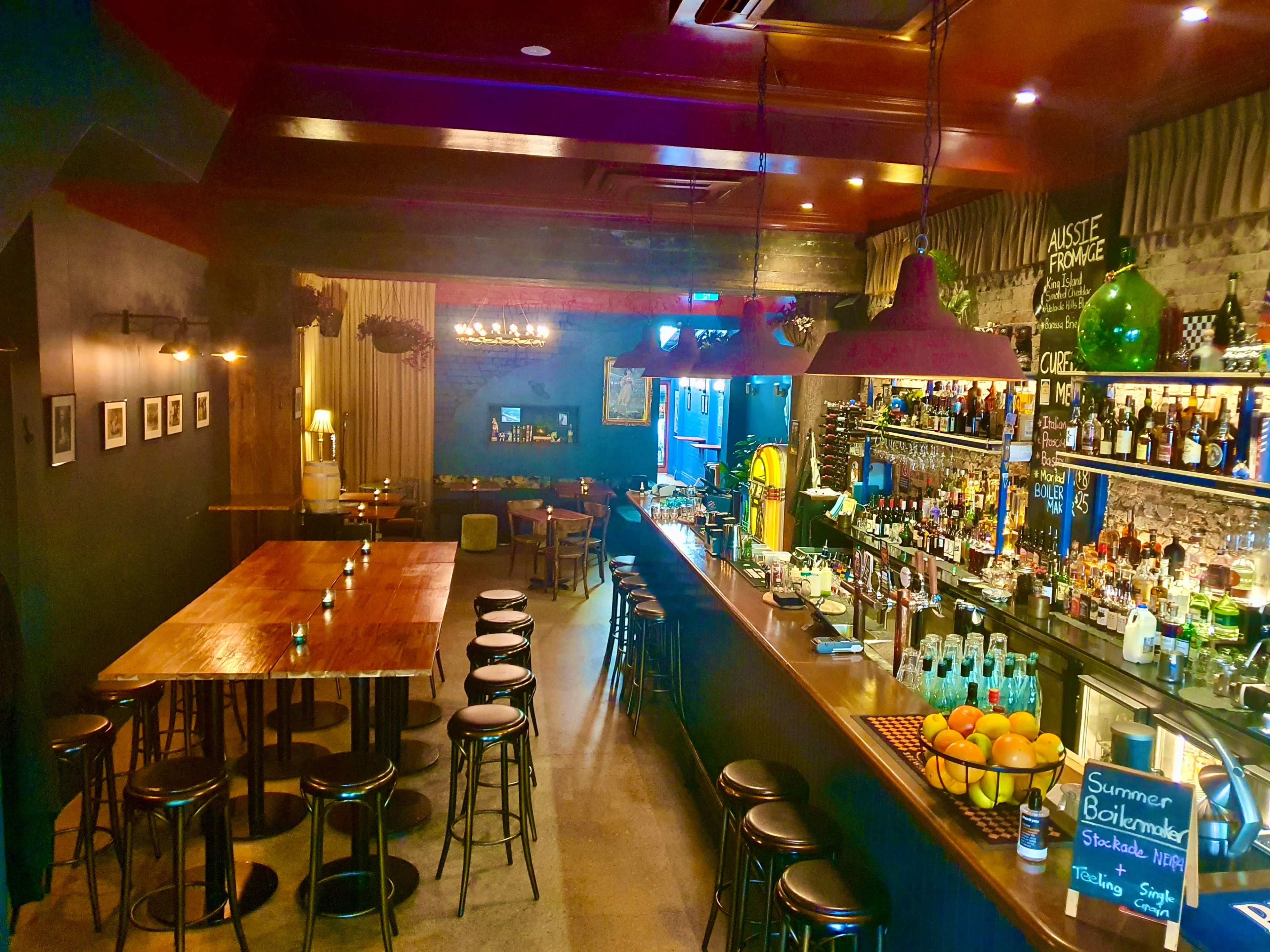 A dimly lit bar with a long counter, high stools, a wooden communal table, and various bottles arranged on shelves behind the bar.