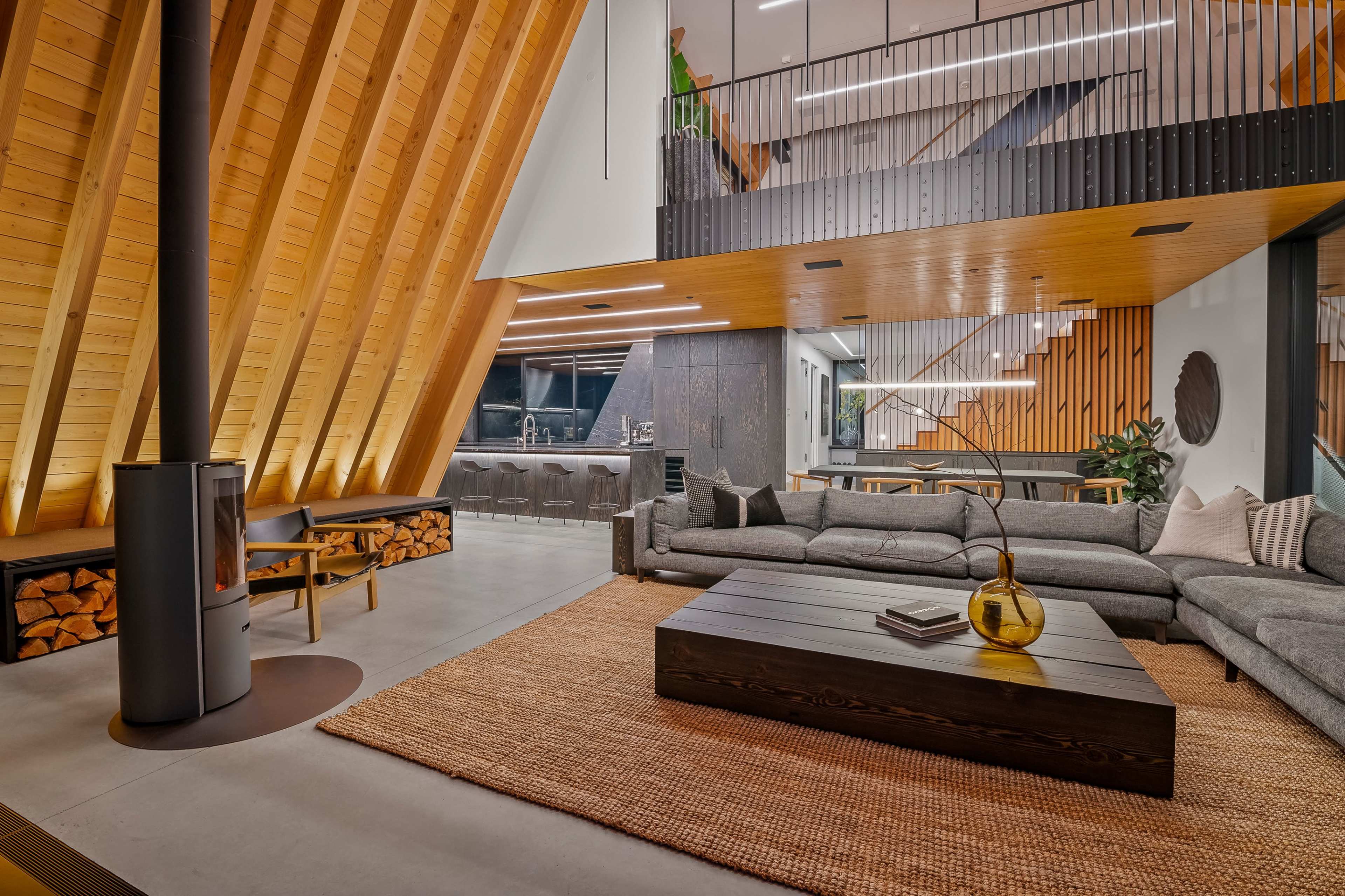 A modern living room features a slanted wooden ceiling, a large sectional sofa, and a central coffee table on a woven rug, with a fireplace and a kitchenette in the background.