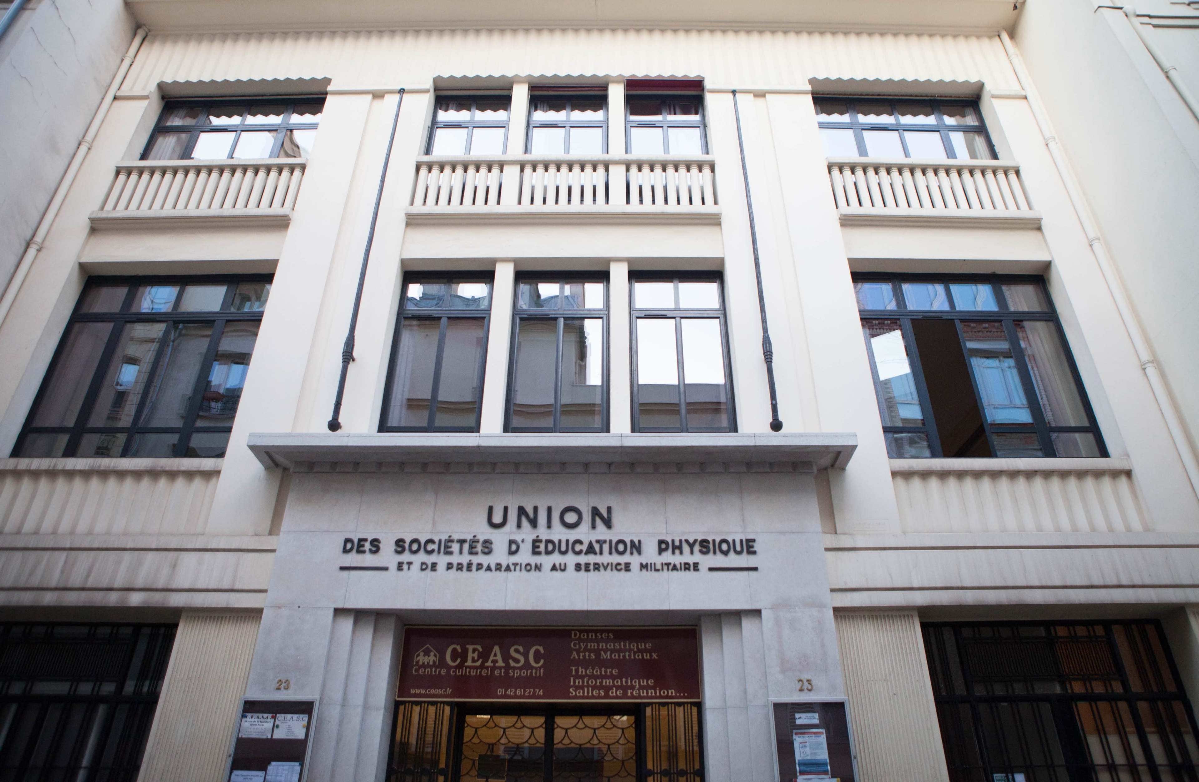 The image shows a historic building with the inscription "UNION DES SOCIÉTÉS D'ÉDUCATION PHYSIQUE ET DE PRÉPARATION AU SERVICE MILITAIRE" prominently displayed above the entrance, featuring large windows and an art deco architectural style.
