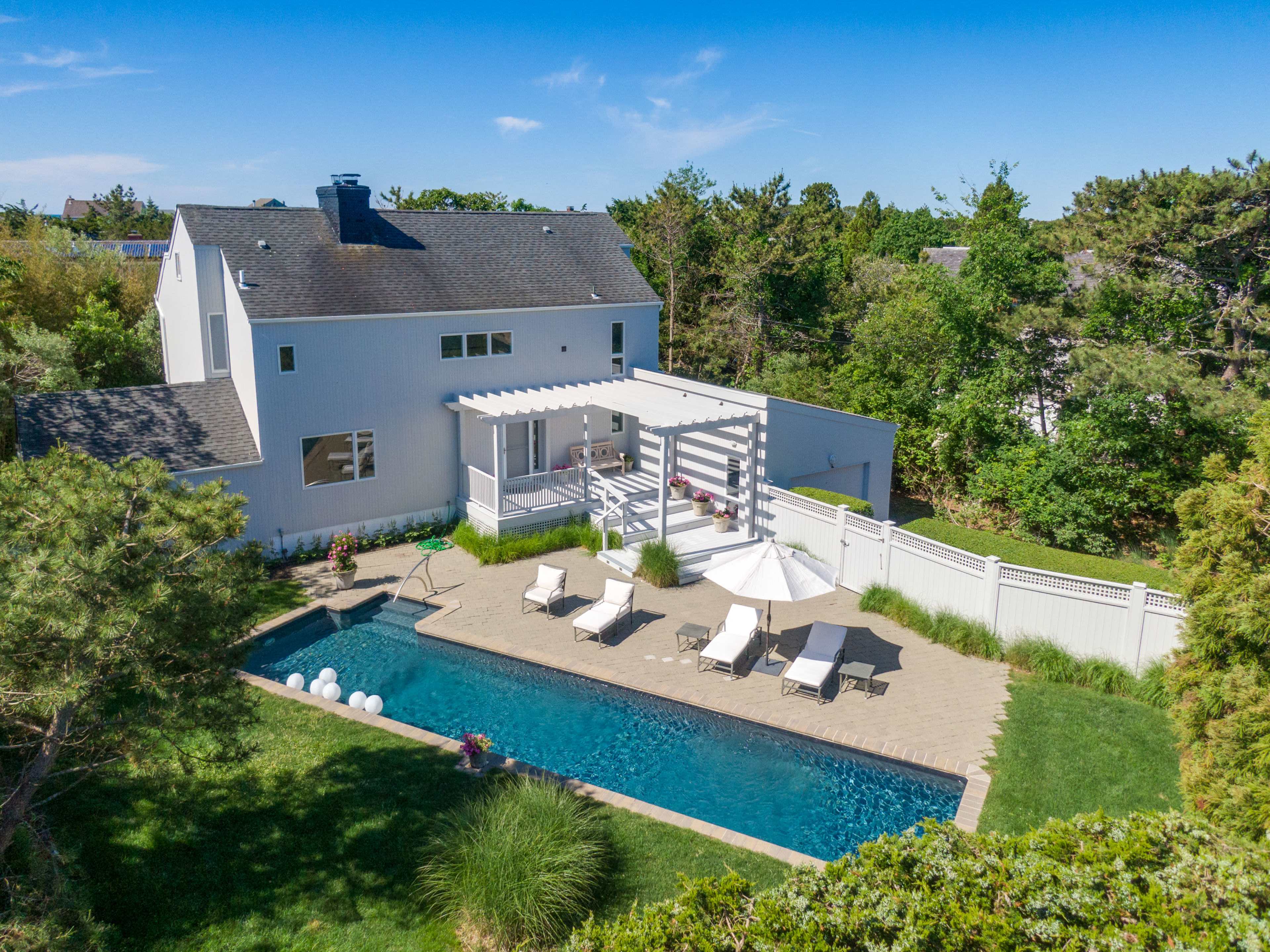 A spacious backyard features a rectangular pool surrounded by lounge chairs and a patio area, with a house and trees in the background.