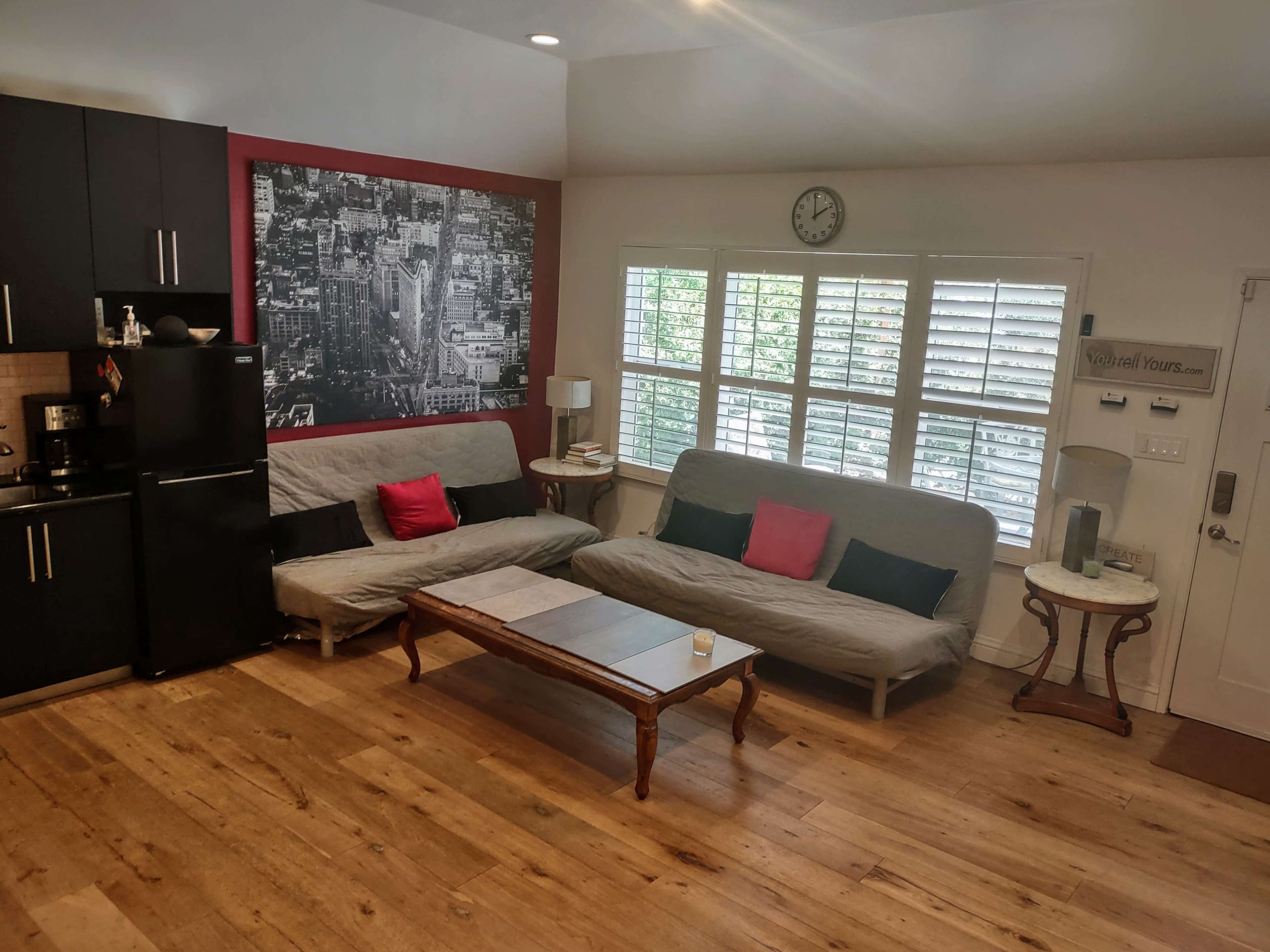 The room features two gray futons with red accent pillows, a wooden coffee table in front, a black refrigerator, and a wall adorned with a black-and-white cityscape.