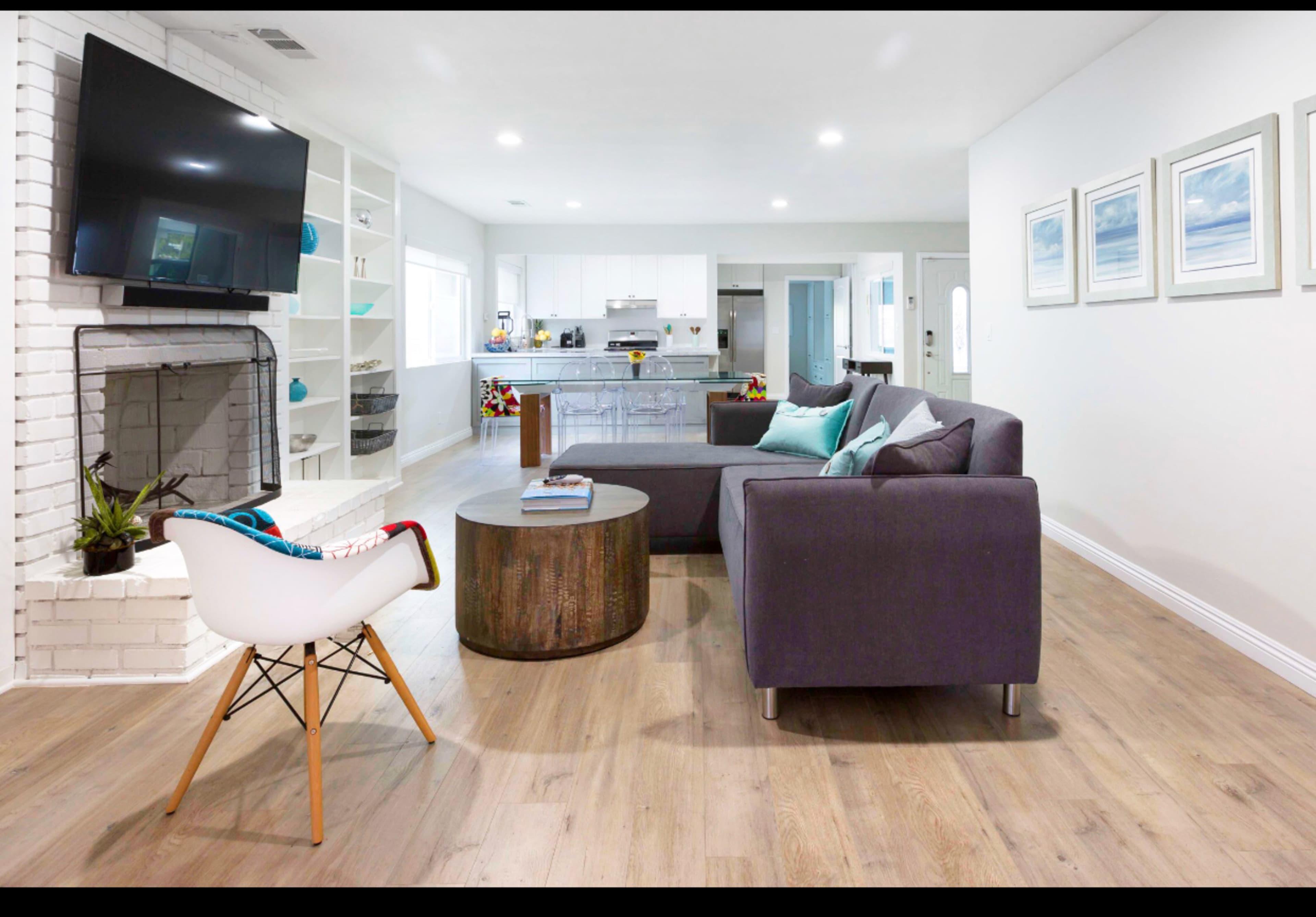 A modern living room with a gray sofa, a round wooden coffee table, and a television mounted on a white brick wall, leading to an open kitchen area.