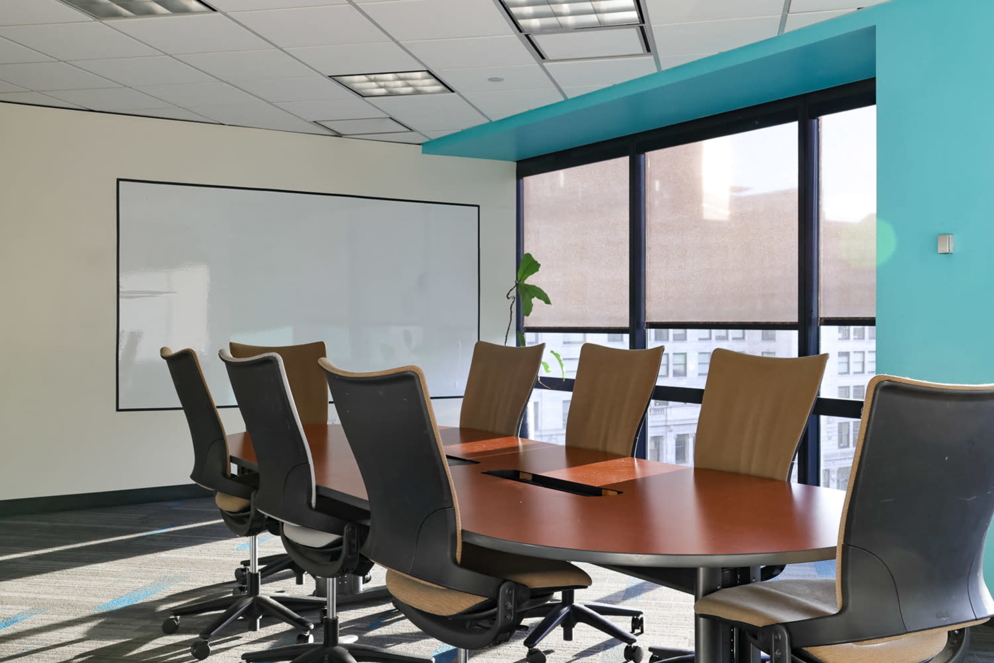 A conference room features a long wooden table with several black office chairs and large windows providing natural light.
