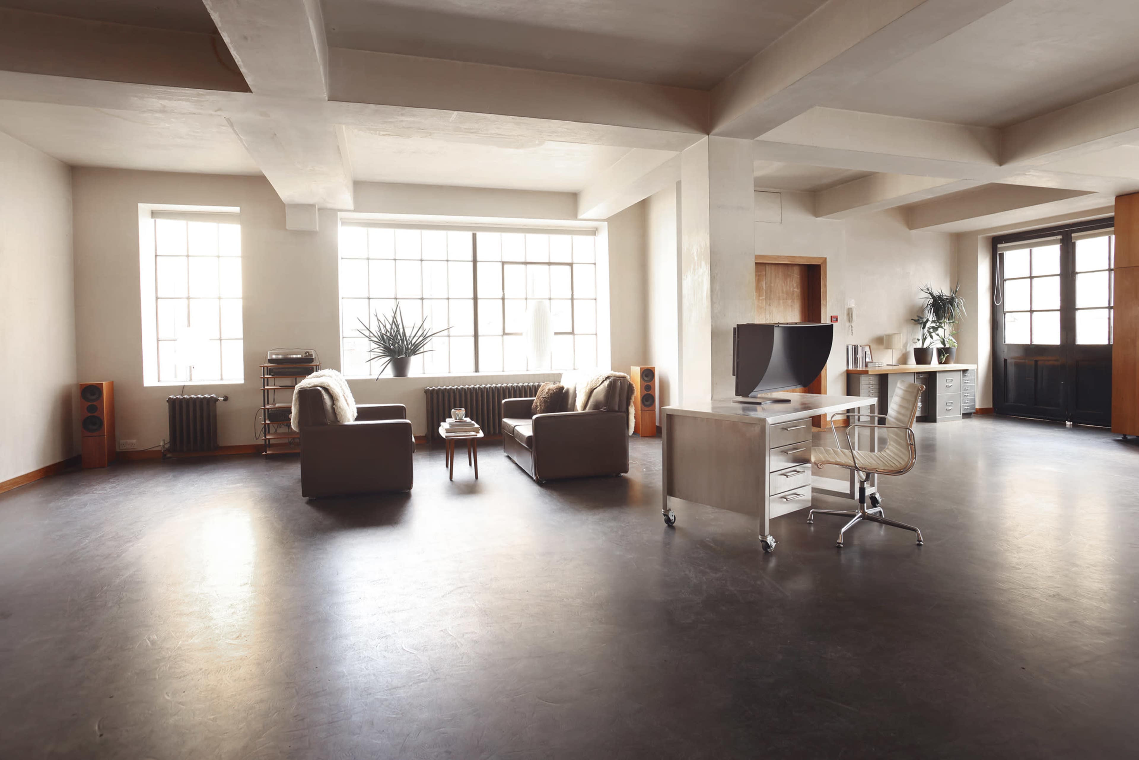 The image depicts a spacious, modern room featuring two sofas, a coffee table, a desk with a chair, and large windows that allow ample natural light.