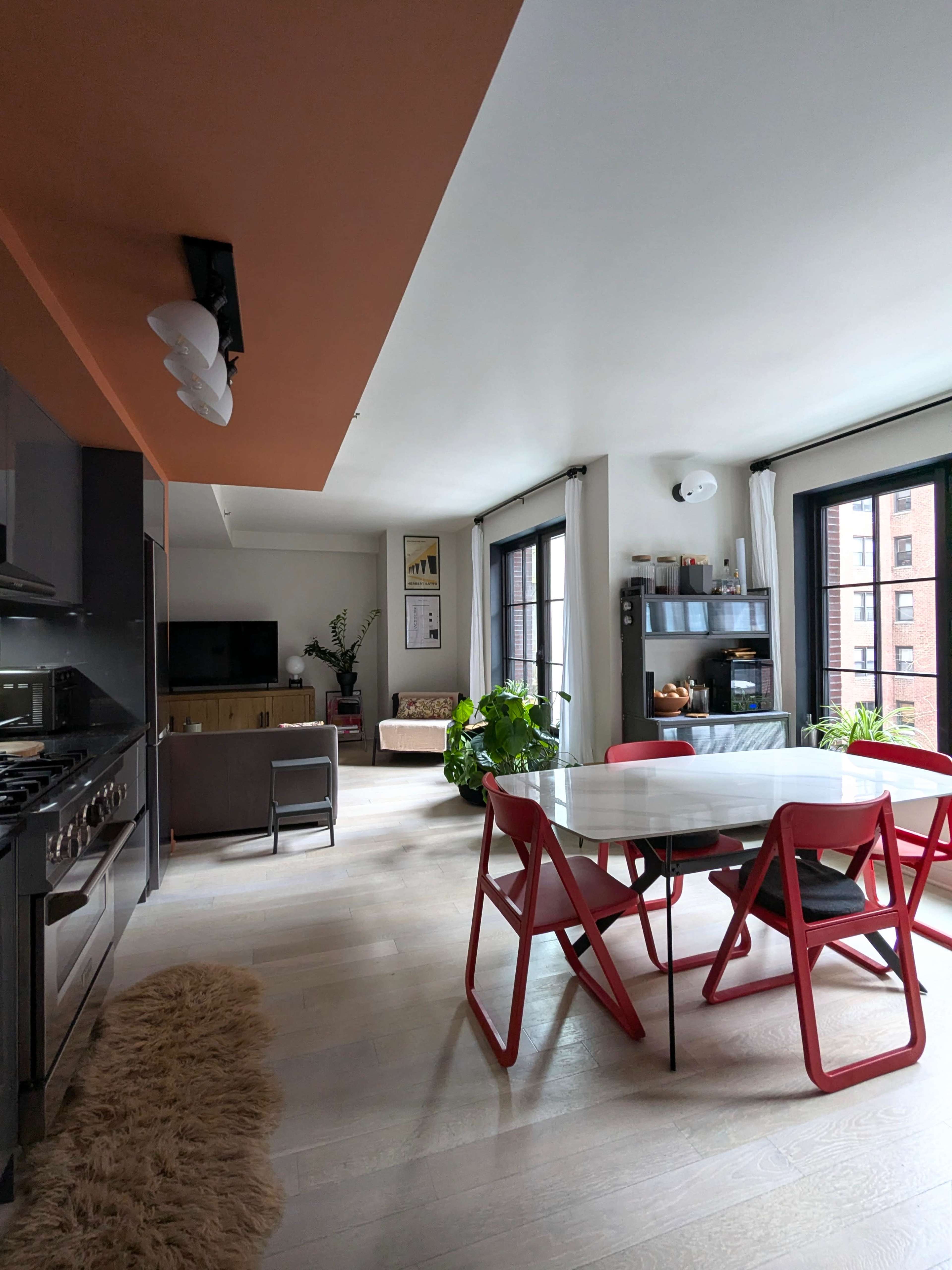 The image shows a modern kitchen and dining area with a dining table and red chairs, a sofa, and large windows allowing natural light to enter the space.