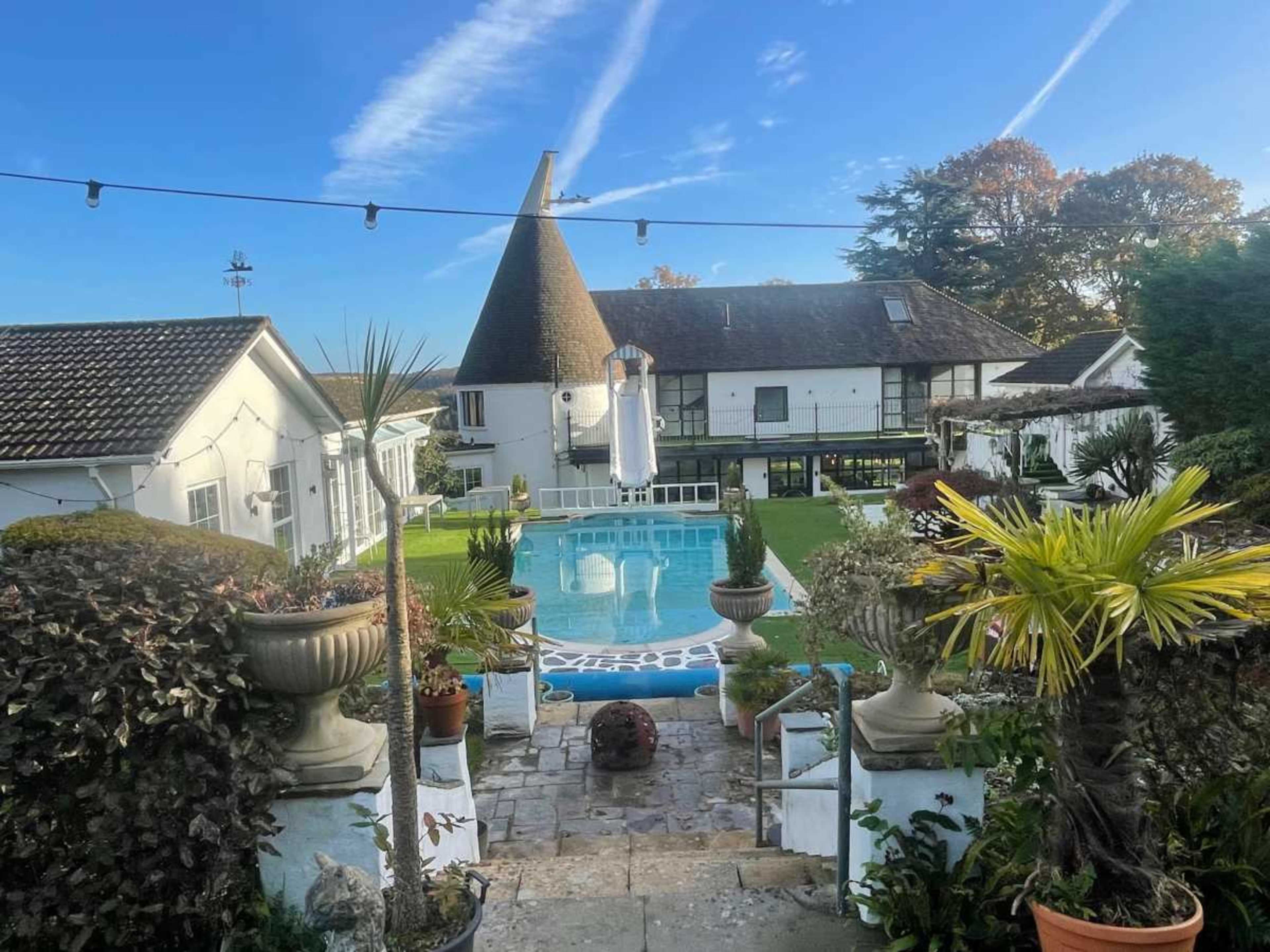 A landscaped garden leading to a swimming pool surrounded by a white building with a distinctive conical roof.