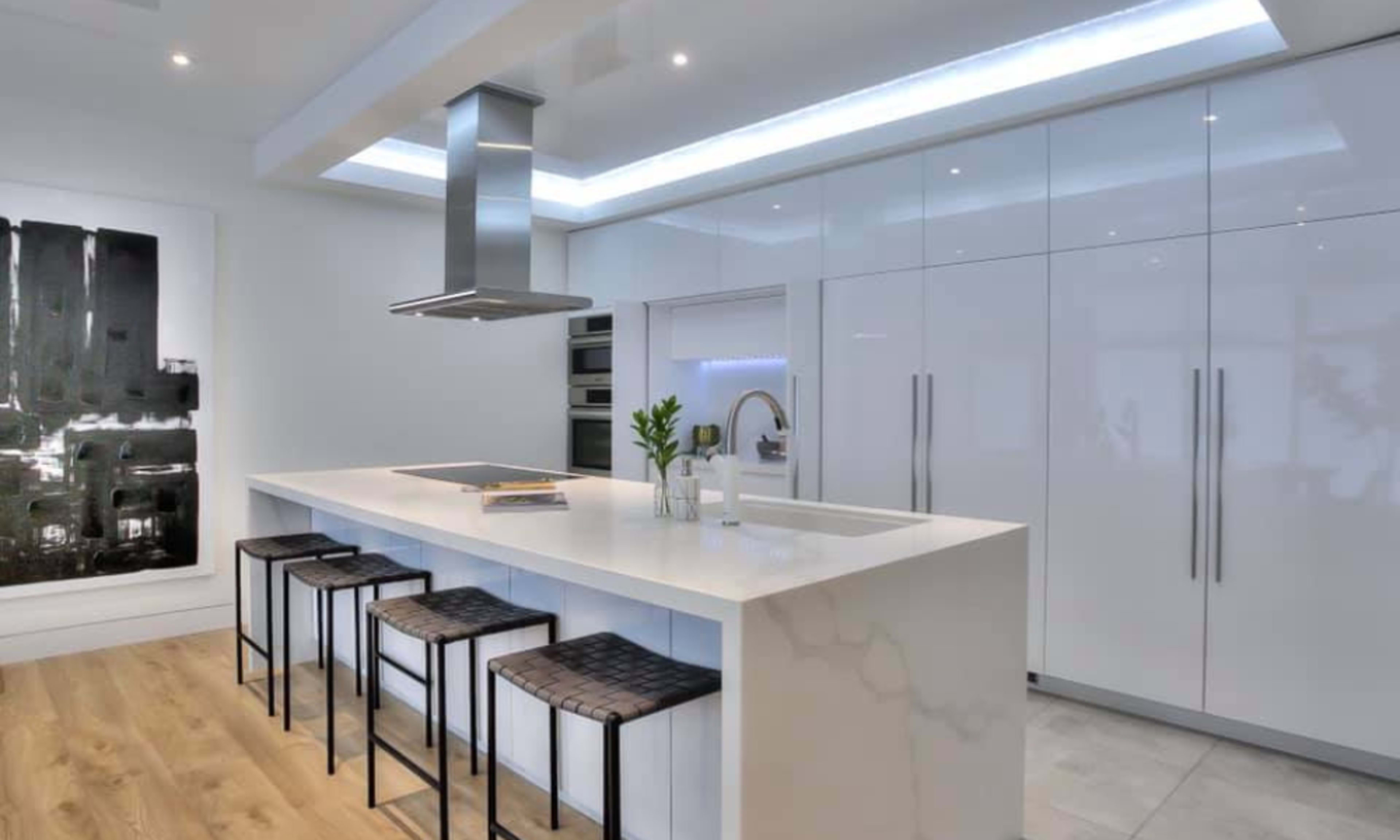 A modern kitchen features a white countertop island with four black stools, sleek cabinetry, and a stainless steel hood above a built-in cooktop.