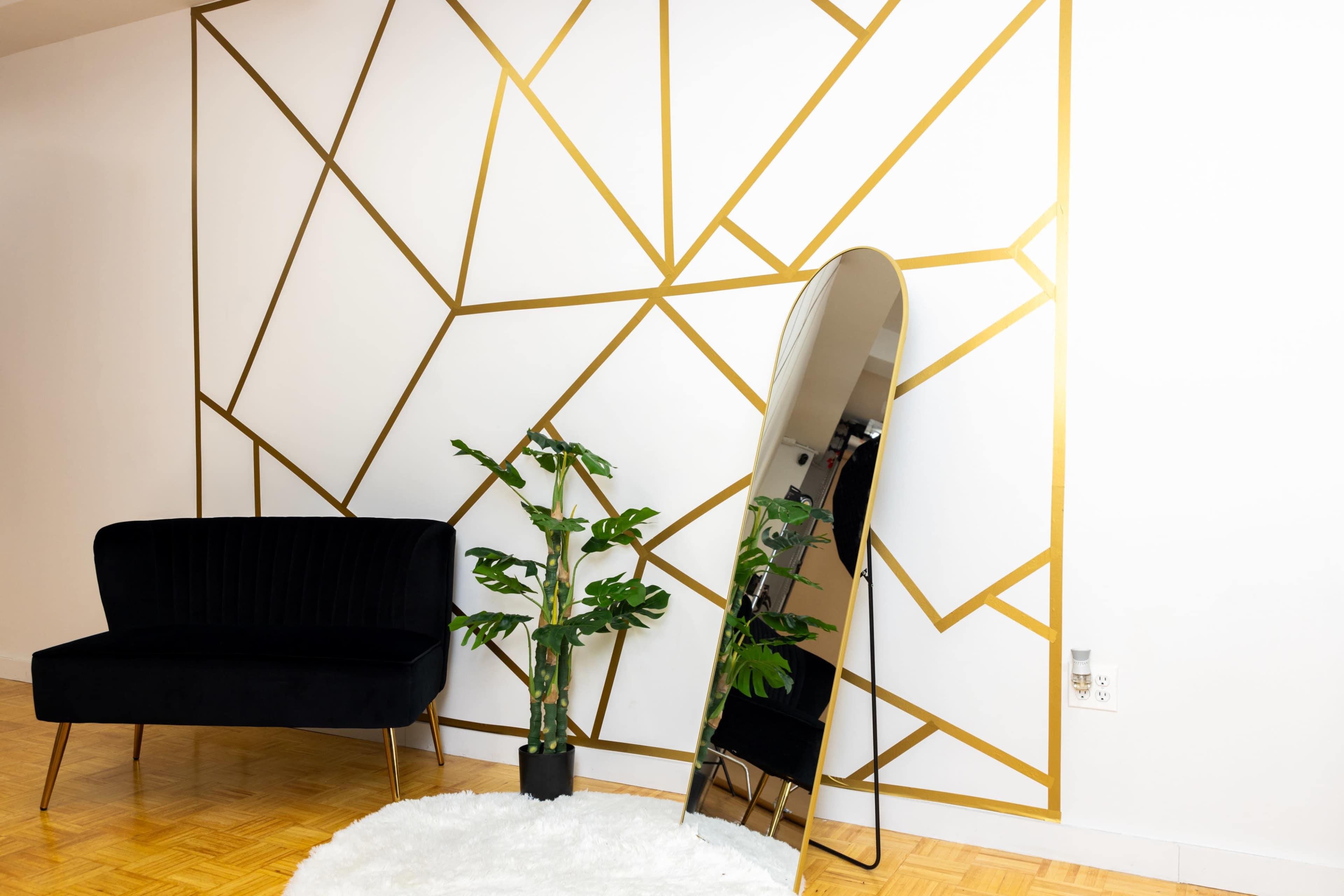 A modern interior with a gold geometric accent wall, a black sofa, a tall mirror, and a potted plant on a circular rug.