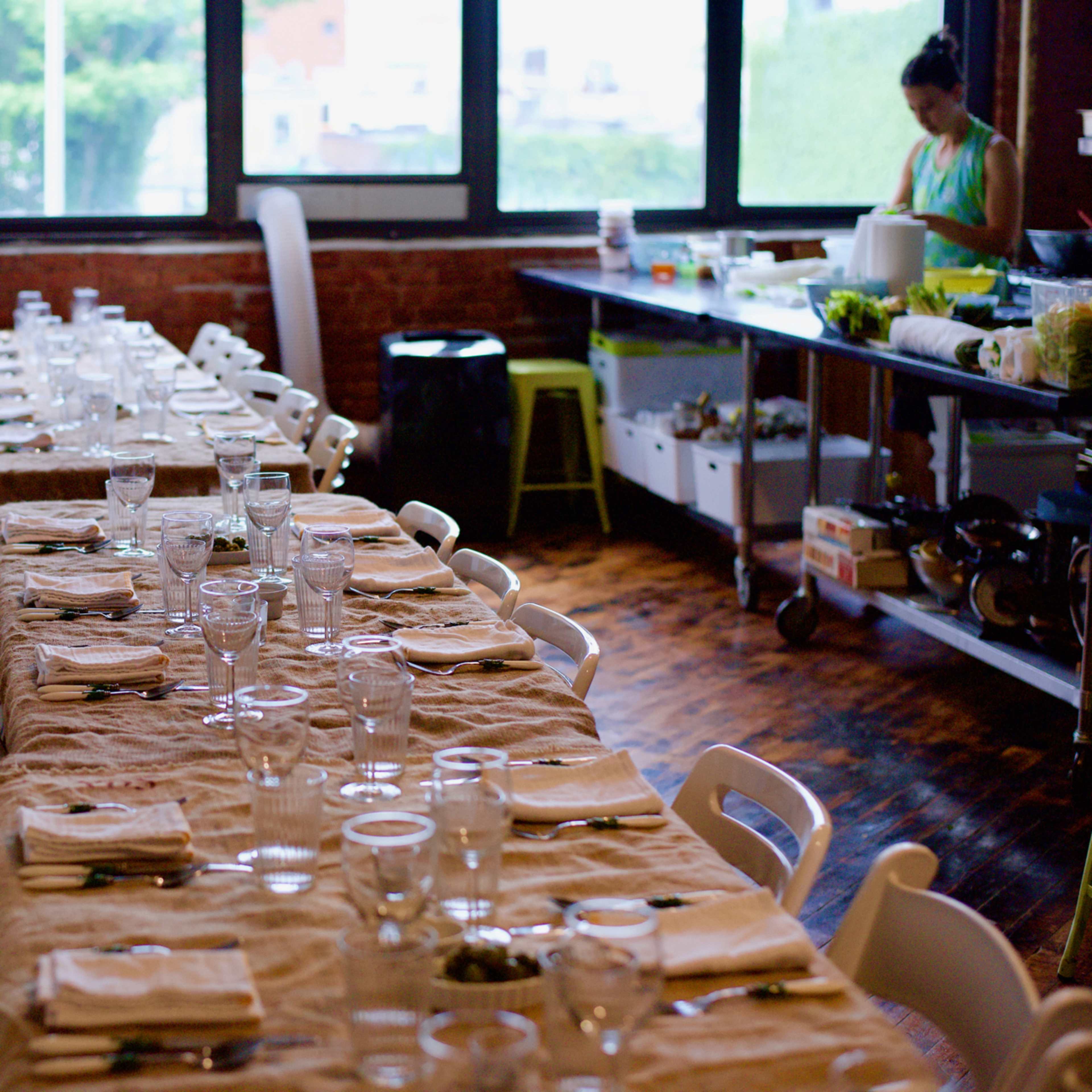 A long dining table is set with clear glasses and white napkins, while a woman prepares food in the background.