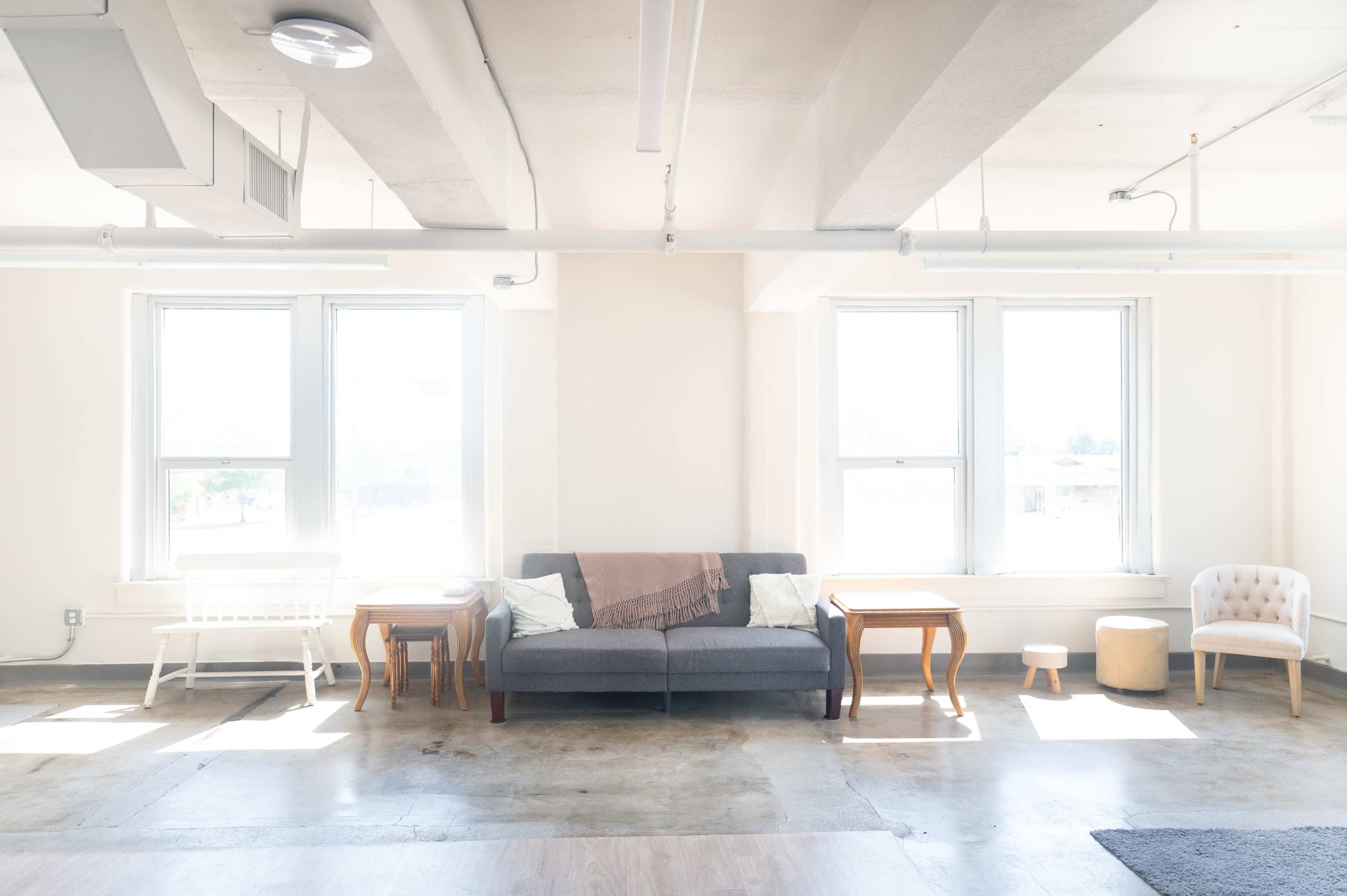 The image shows a bright, minimalistic room with a couch, two small tables, a pouf, and a single armchair by the windows.