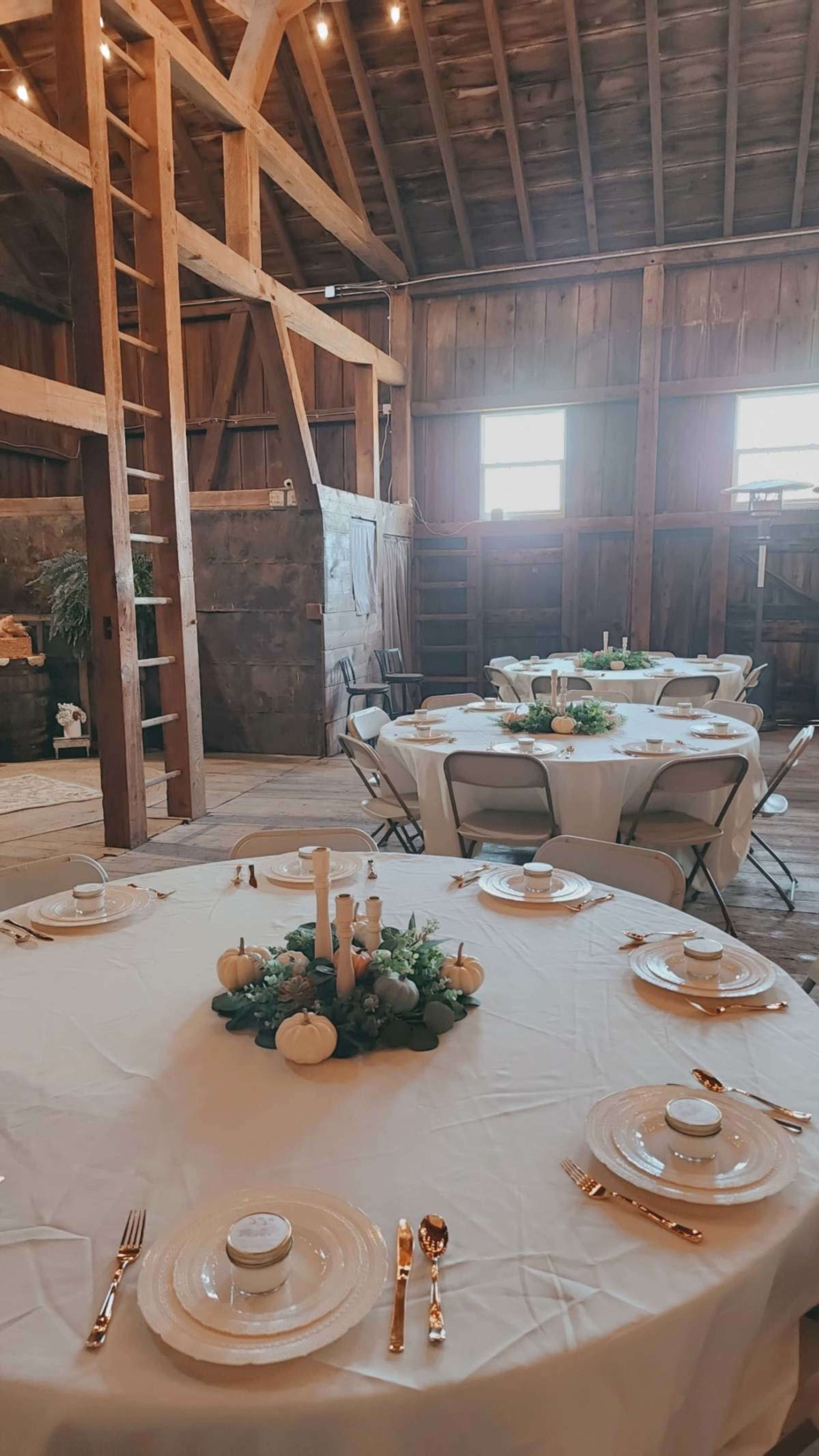 The interior of a rustic barn features a table set for an event, complete with plates, utensils, and decorative elements, surrounded by wooden beams and dim lighting.
