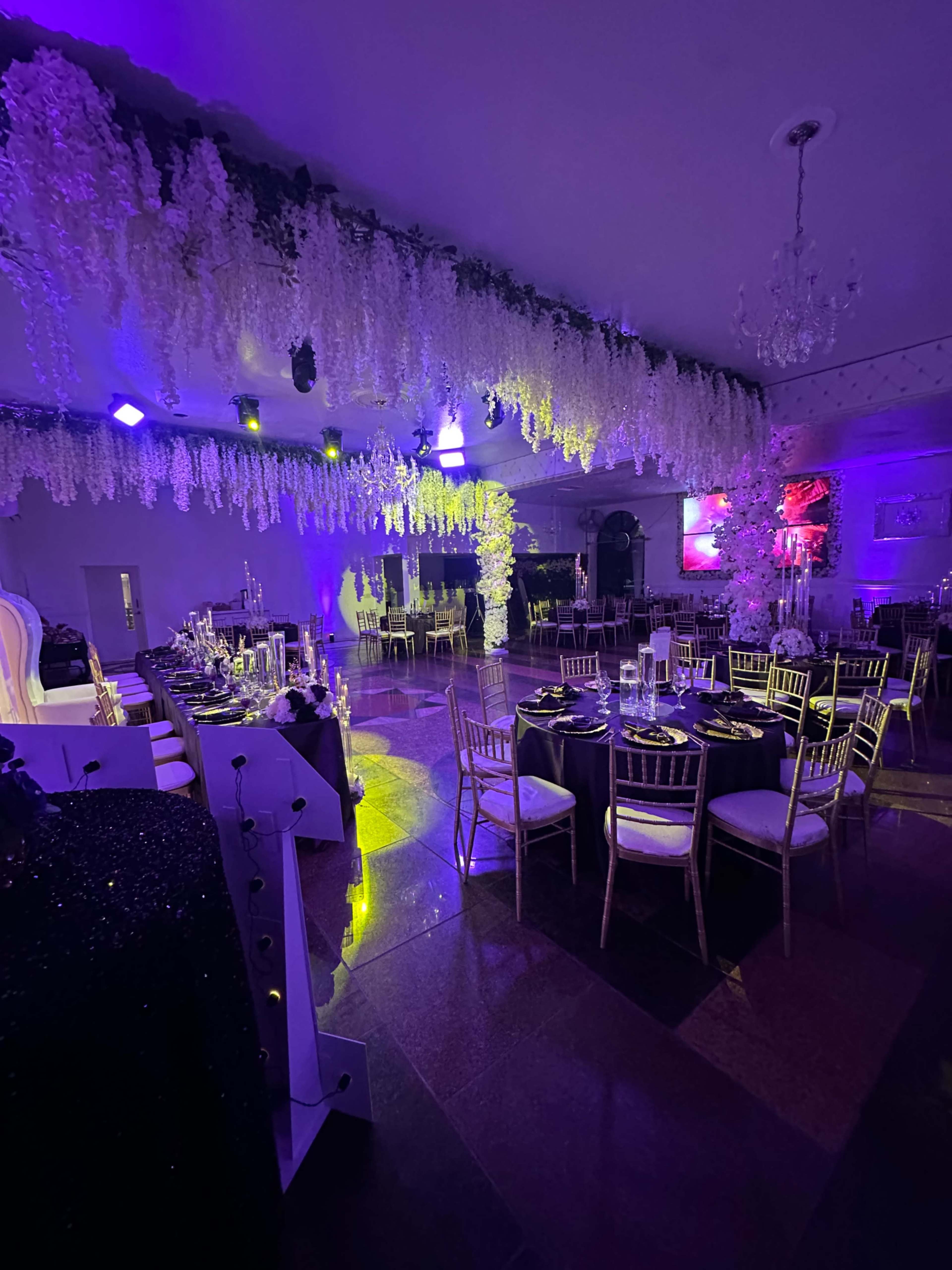 The image shows a decorated banquet hall with elegant tables set for a formal event, featuring purple lighting and floral arrangements hanging from the ceiling.