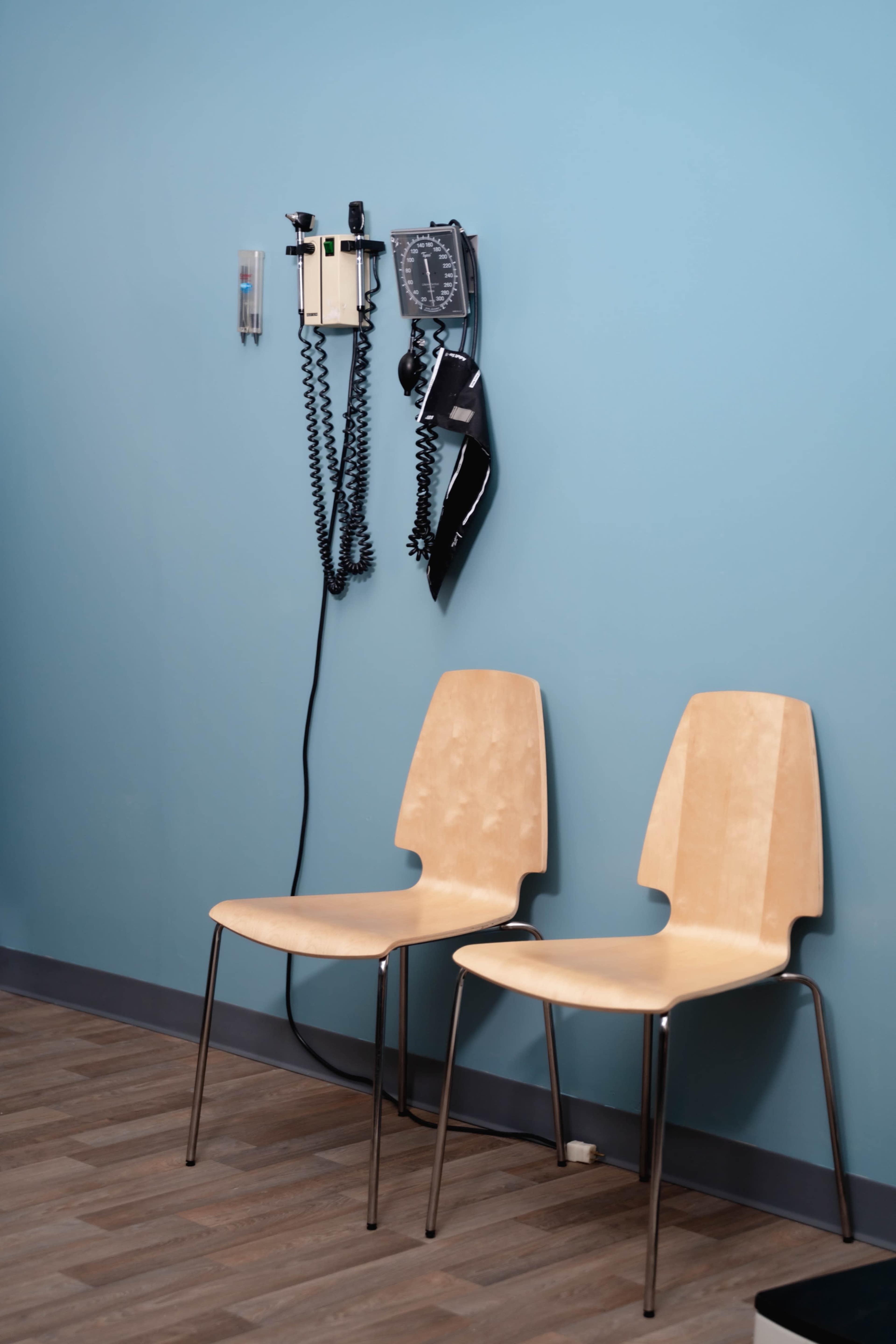 Two light-colored chairs are positioned against a blue wall, with medical equipment and a sphygmomanometer hanging nearby.