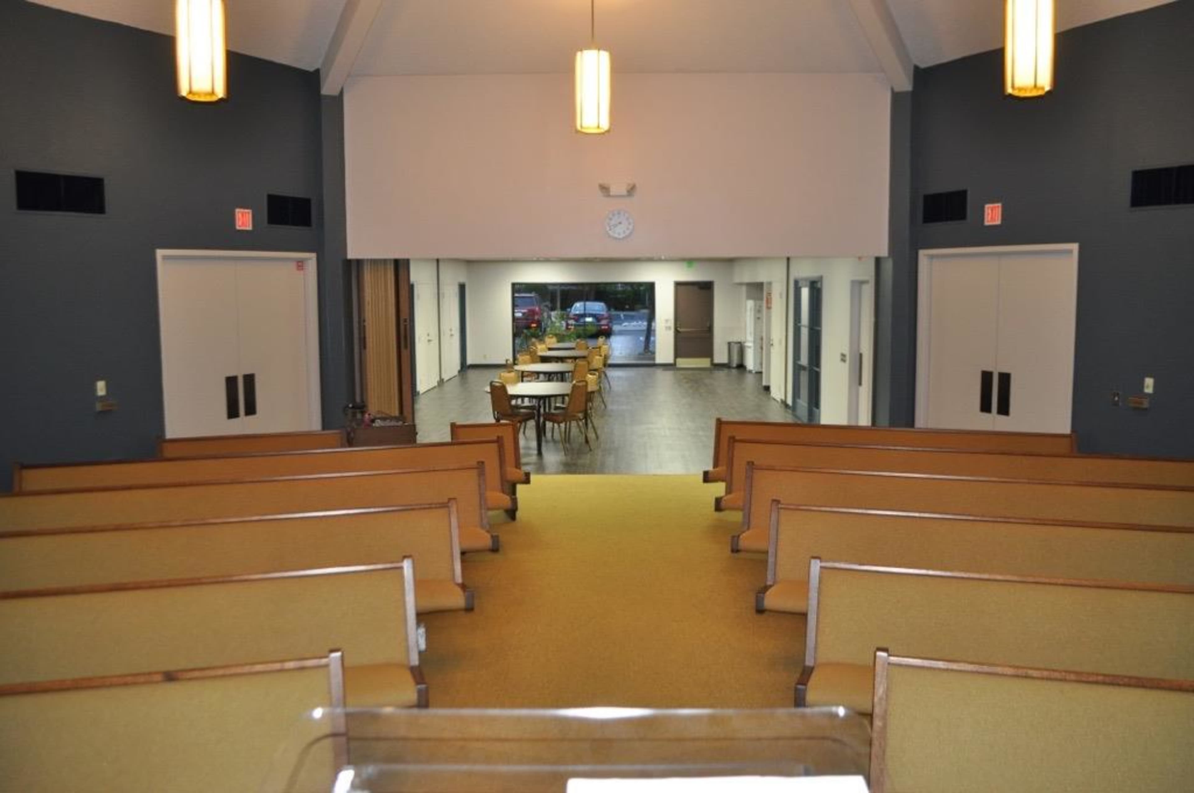 An empty church interior with rows of wooden pews facing a large open area containing tables and chairs.