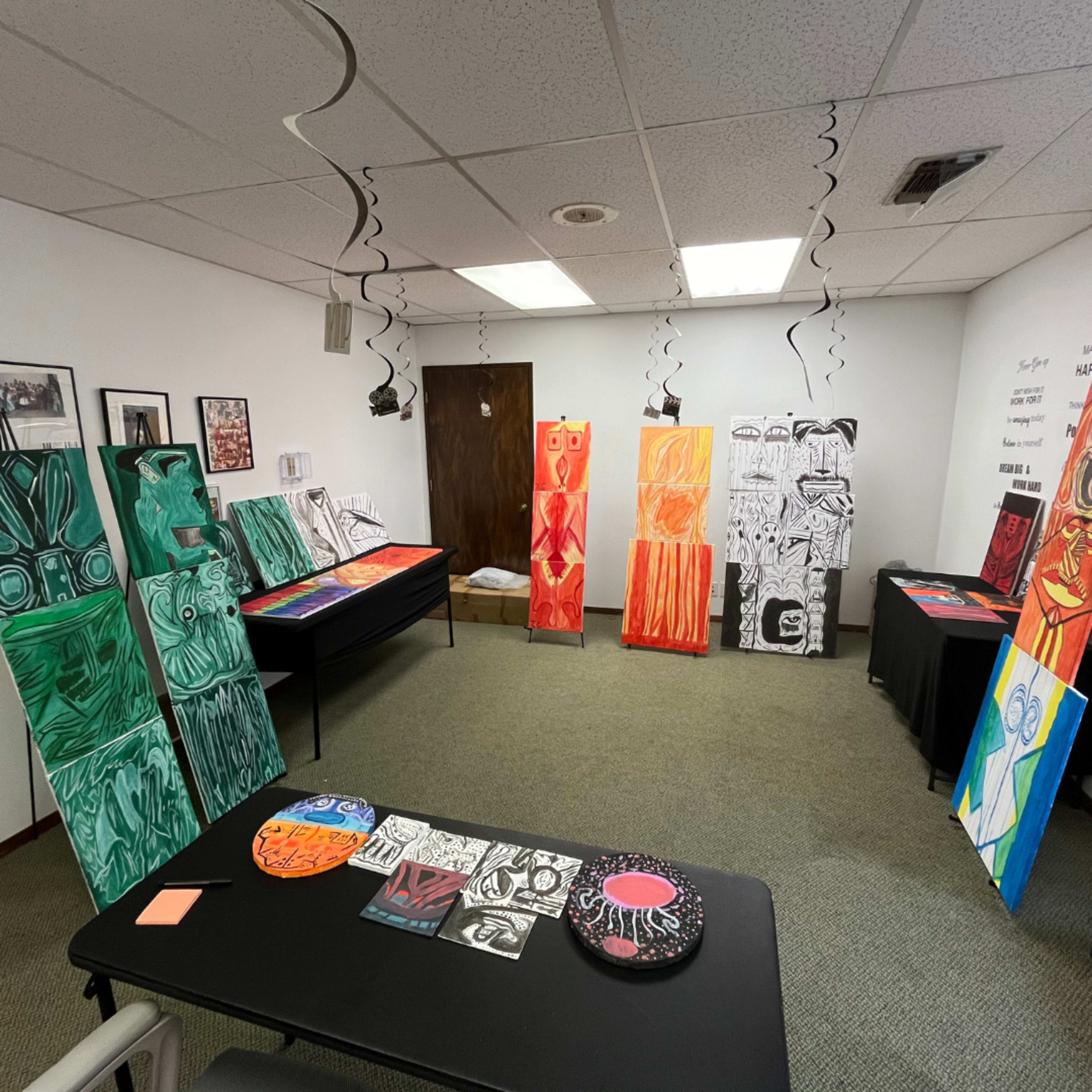 The room features several large and colorful paintings displayed on easels and tables, with black and white streamers hanging from the ceiling.