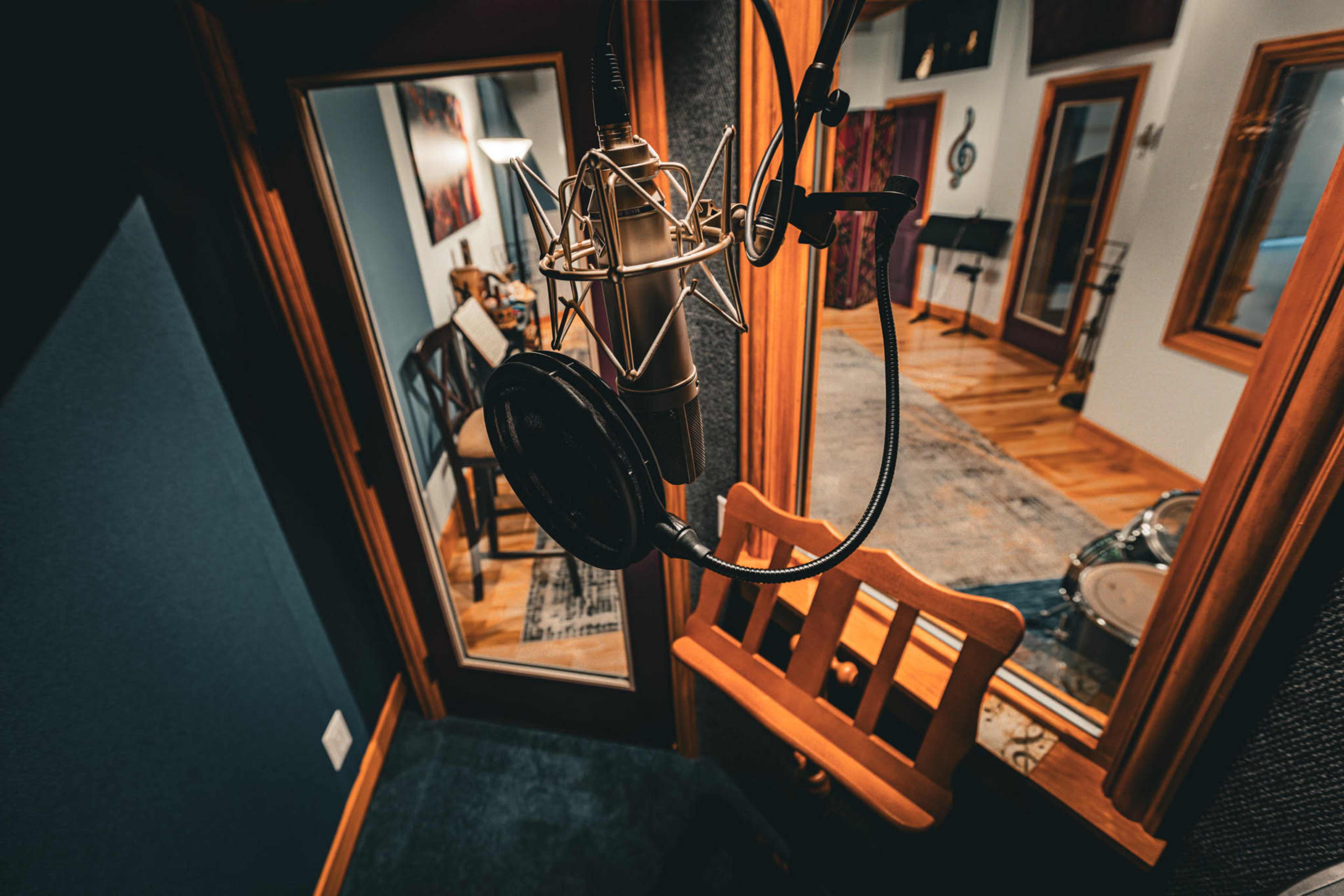 The image shows a recording studio featuring a microphone with a pop filter and a chair, framed by glass windows overlooking an instrument area.